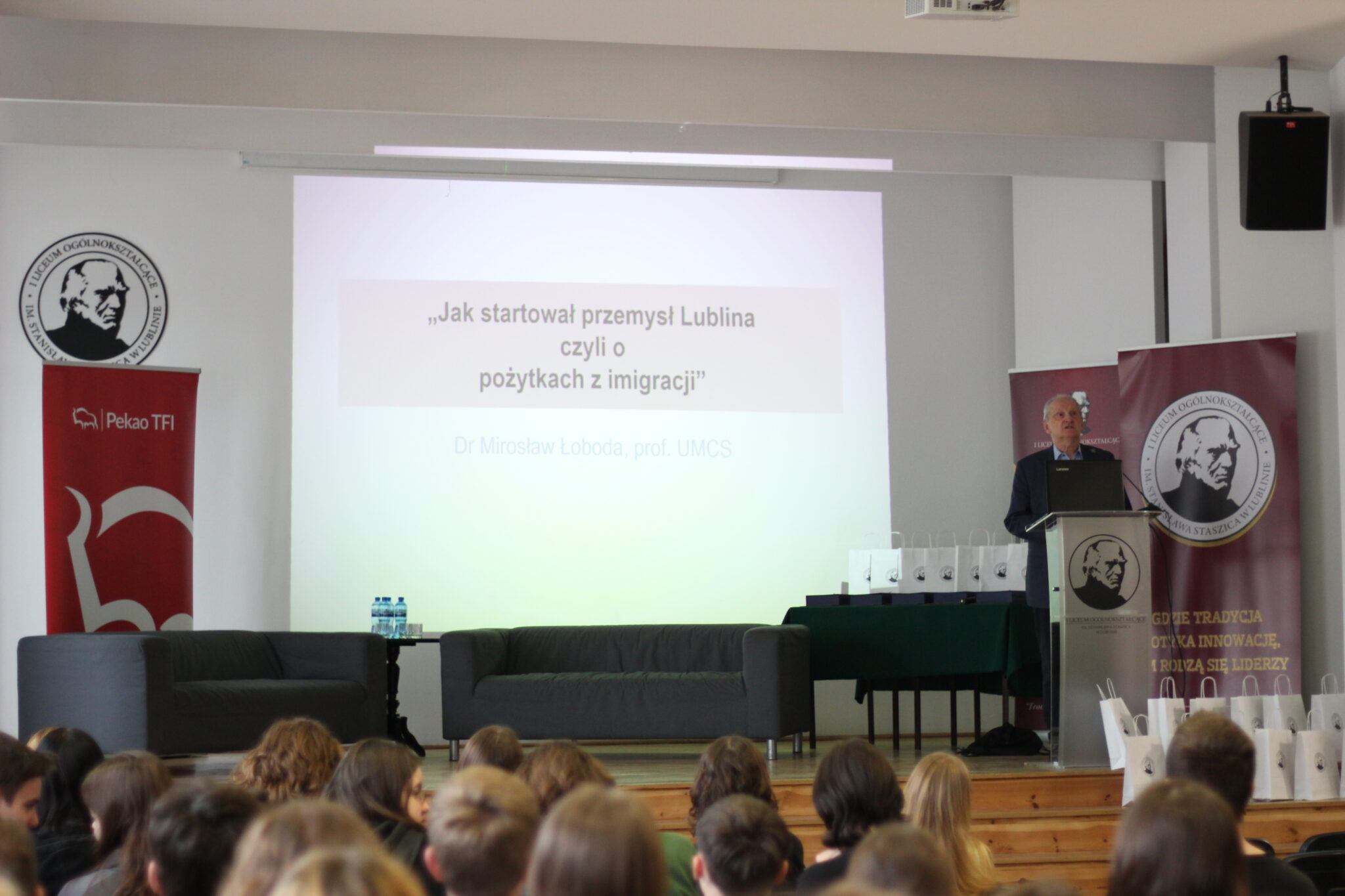 Zdjęcie przedstawia moment wręczenia nagrody podczas wydarzenia edukacyjnego „I Młodzieżowe Forum Ekonomiczne – Staszic 2024”. W centrum kadru znajdują się trzy osoby pozujące do wspólnej fotografii – mężczyzna i dwie młode kobiety. Mężczyzna trzyma dyplom z nagrodą. W tle widoczny jest duży ekran z rzutnikiem, prezentujący informacje o nagrodzie i jej laureacie. Scena znajduje się w szkolnej sali konferencyjnej. Szczegółowy opis postaci i ustawienia: Osoby: Środek kadru: Dorosły mężczyzna w średnim wieku, siwiejący, z krótkim zarostem i okularami. Ubrany w elegancką marynarkę w granatową kratę i jasnoniebieską koszulę. Trzyma w dłoniach przezroczystą ramkę lub oprawiony dyplom z tekstem i logiem szkoły. Po jego lewej stronie (prawa strona zdjęcia): Młoda kobieta z długimi, ciemnymi włosami, ubrana w czarną marynarkę i jasny top. Uśmiecha się lekko i stoi z rękami wzdłuż ciała. Po jego prawej stronie (lewa strona zdjęcia): Młoda kobieta z długimi brązowymi włosami, ubrana w czarne spodnie i czarny, dzianinowy bezrękawnik z białą koszulą z kołnierzem pod spodem. Również uśmiechnięta, ma ręce schowane za plecami. Tło: W tle znajduje się ekran projekcyjny z wyświetlonym slajdem. Slajd zawiera tekst i grafikę: Nagłówek: „Nagroda w kategorii SUKCES W DZIAŁALNOŚCI SPOŁECZNEJ NA RZECZ MŁODZIEŻY” (część w czerwonym kolorze). Opis: „Program Horyzonty Fundacji im. prof. Romana Czarneckiego” Dalej: „Pan Igor Brewka pełniący obowiązki Prezesa Fundacji” – mężczyzna na zdjęciu to najprawdopodobniej pan Igor Brewka. W prawym górnym rogu slajdu umieszczono logo I LO im. Staszica (okrągłe, czarno-białe z profilem mężczyzny) oraz czerwony sześciokąt z trójwymiarową, srebrną strzałką skierowaną w górę i prawo. Na samym dole ekranu – częściowo widoczny napis: „I MŁODZIEŻOWE FORUM EKONOMICZNE STASZIC 2024”. Scenografia sali: Przed osobami na zdjęciu stoją dwa ciemnoszare fotele i mały czarny stolik z sześcioma butelkami wody mineralnej. Po prawej stronie kadru widoczny ciemnozielony obrus przykrywający stół lub mównicę, częściowo poza kadrem. Podłoże jasne, scena dobrze oświetlona światłem sztucznym. Znaczenie zdjęcia: Fotografia dokumentuje uroczysty moment wręczenia nagrody Fundacji Horyzonty w ramach forum ekonomicznego dla młodzieży. Nagroda dotyczy działań społecznych na rzecz młodych ludzi. Zdjęcie podkreśla współpracę między instytucjami edukacyjnymi a fundacjami wspierającymi rozwój młodzieży.