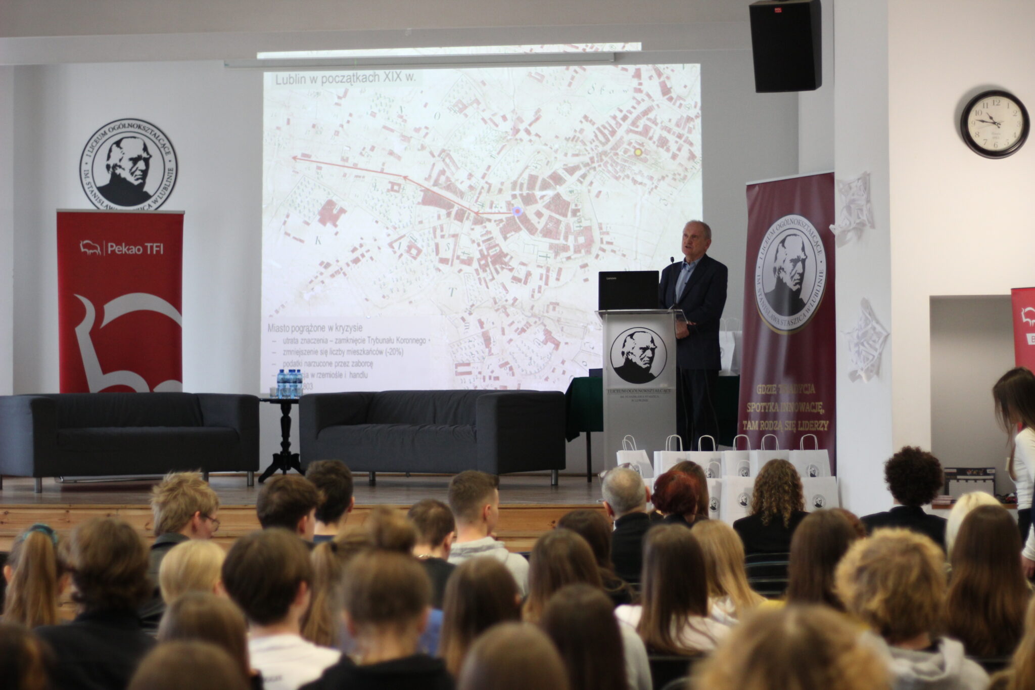 To samo miejsce i prelegent. Na ekranie szczegółowa, zabytkowa mapa Lublina z początku XIX wieku, z podpisem „Miasto pogrążone w kryzysie” oraz punktami: utrata znaczenia, spadek liczby mieszkańców, podatki narzucone przez zaborcę. Prelegent kontynuuje wykład przy mównicy. W tle znane już banery, obok widoczna tarcza zegara na ścianie.