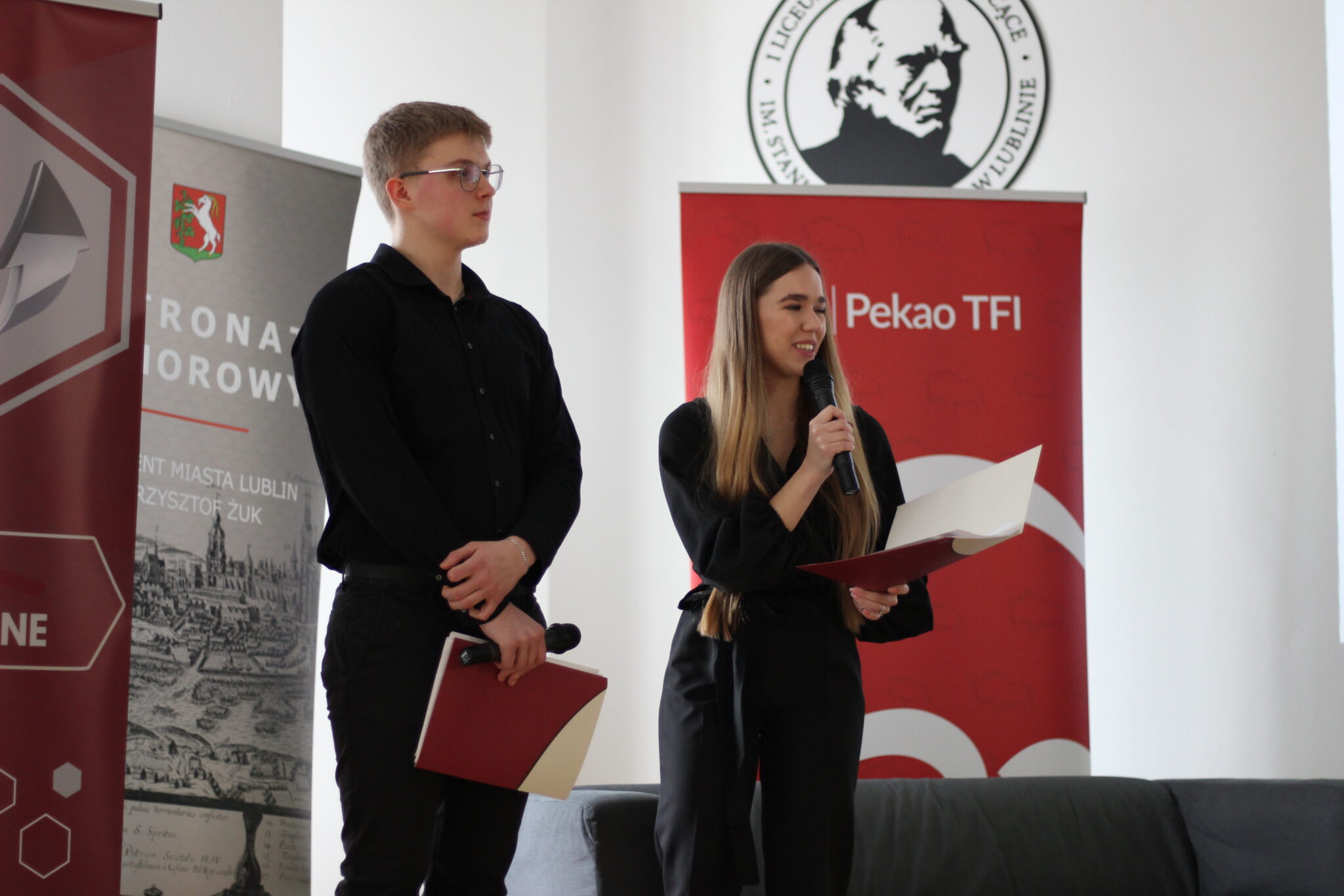 Dwoje młodych ludzi – chłopak i dziewczyna w czarnych eleganckich ubraniach – stoi na scenie z mikrofonami i bordowymi teczkami. Dziewczyna przemawia z uśmiechem, chłopak trzyma mikrofon i patrzy przed siebie. Za nimi banery szkoły i miasta Lublin, w tym herb Lublina oraz logotyp Pekao TFI.