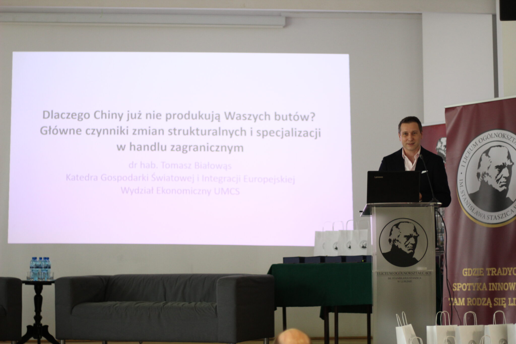 Mężczyzna w garniturze i jasnoróżowej koszuli stoi za mównicą. Na ekranie slajd z tytułem wykładu: „Dlaczego Chiny już nie produkują Waszych butów? Główne czynniki zmian strukturalnych i specjalizacji w handlu zagranicznym”. Pod spodem podpis: dr hab. Tomasz Białowąs, Katedra Gospodarki Światowej i Integracji Europejskiej, UMCS. W tle znane elementy scenografii.