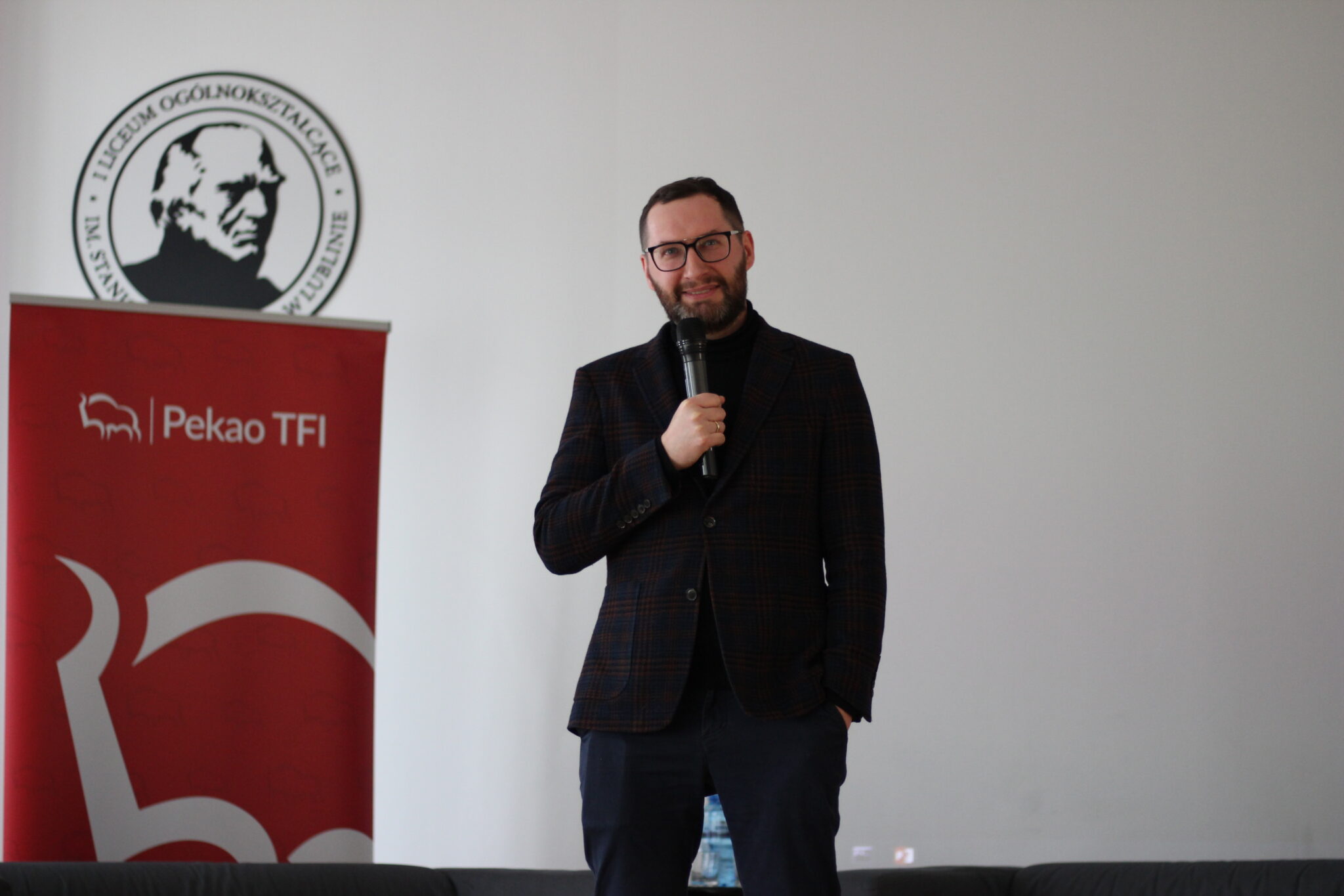 Mężczyzna z brodą i okularami, ubrany w ciemny garnitur w kratę i czarny golf, stoi z mikrofonem, patrząc z uśmiechem w stronę widowni. Za nim czerwony baner Pekao TFI i logo szkoły na ścianie.