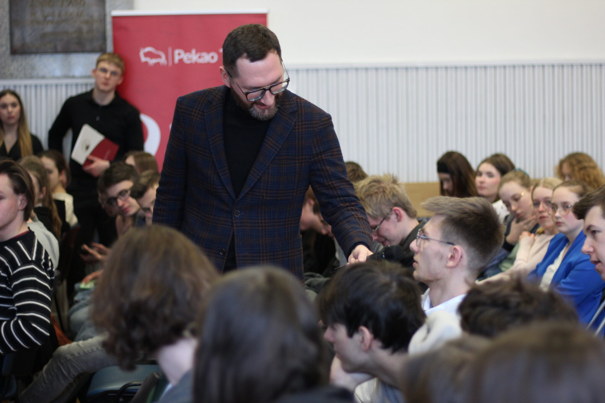 Mężczyzna z brodą, w okularach i granatowej marynarce w kratę pochyla się w stronę młodego uczestnika siedzącego wśród publiczności. Trzyma mikrofon i podaje go uczniowi, który najprawdopodobniej chce zadać pytanie. W tle liczna grupa młodzieży i charakterystyczny czerwony baner z logotypem „Pekao TFI”.