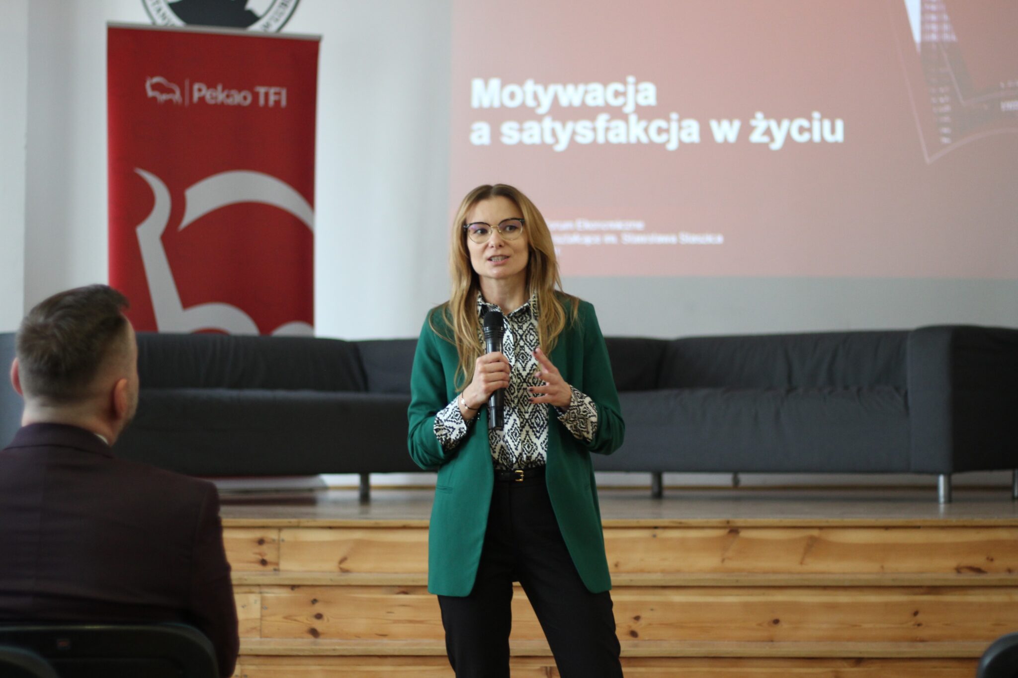 Młoda kobieta z długimi, jasnymi włosami i okularami prowadzi wykład. Ubrana jest w zieloną marynarkę i elegancką bluzkę w biało-czarny wzór. Trzyma mikrofon i przemawia do publiczności. Za nią widoczny slajd z tytułem prezentacji: „Motywacja a satysfakcja w życiu” oraz baner „Pekao TFI”.