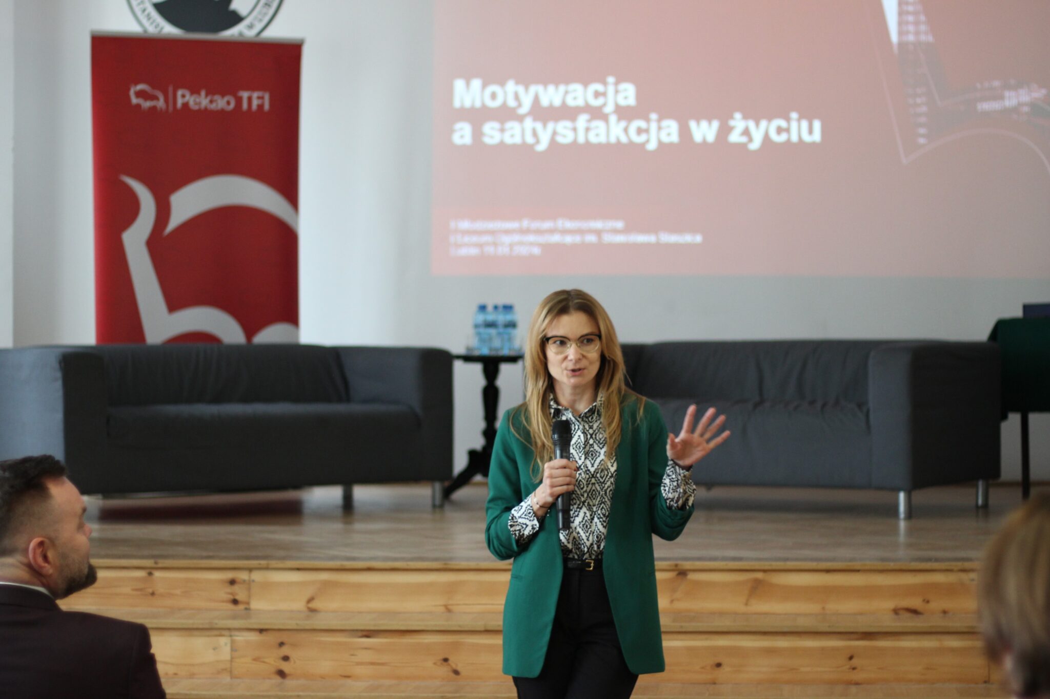 Ta sama prelegentka co wcześniej. Mówi i gestykuluje, mając przed sobą siedzącego mężczyznę w bordowej marynarce (plecami do aparatu). W tle ten sam czerwony slajd z tytułem oraz scenografia sceny szkolnej.