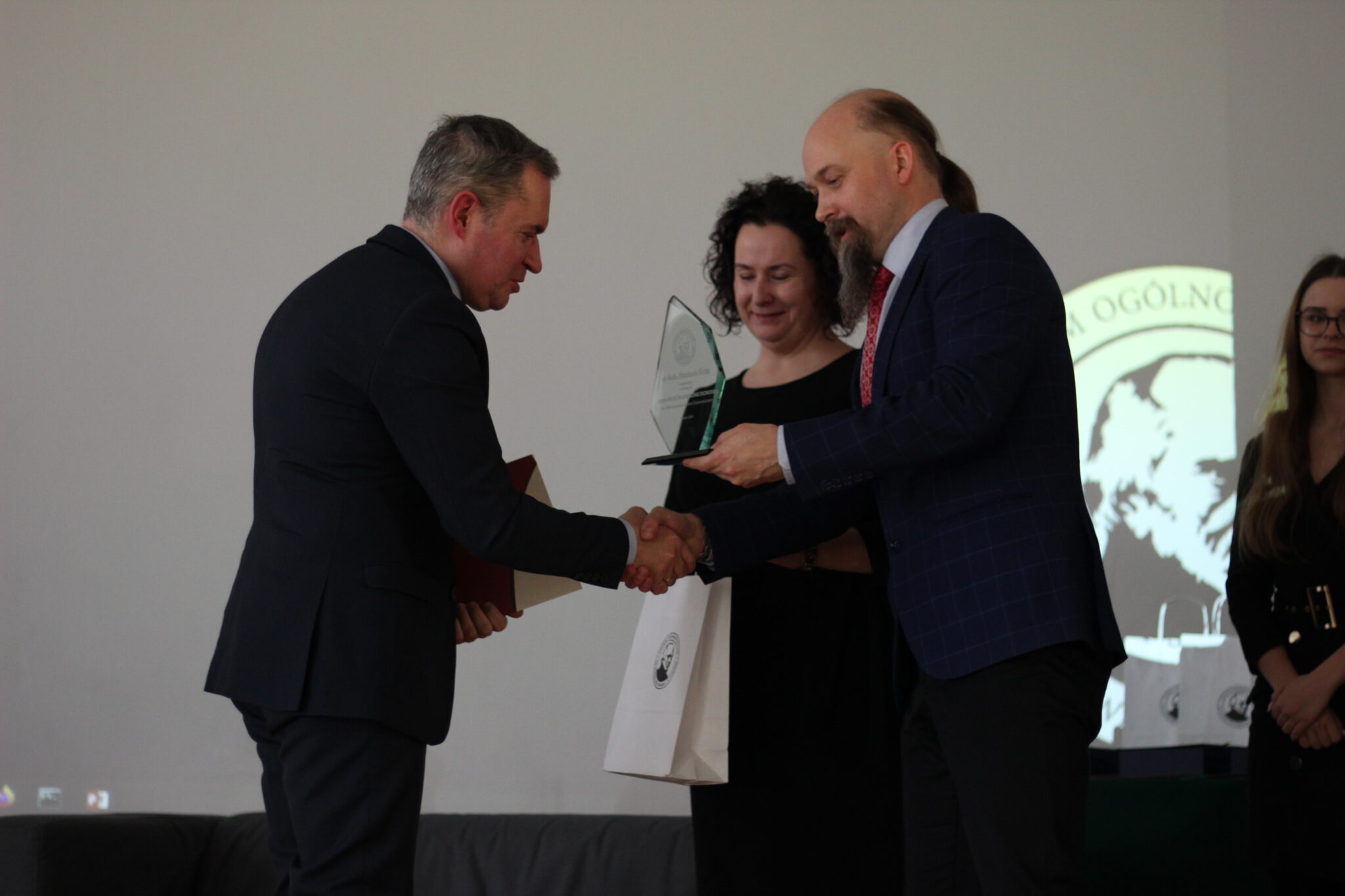 Mężczyzna z siwymi włosami i ciemnym garniturem otrzymuje nagrodę – przezroczystą statuetkę i białą torbę – od brodatego prowadzącego w granatowej marynarce. Mężczyźni ściskają sobie dłonie, kobieta w czarnej sukni stoi obok i uśmiecha się. W tle widoczne jest logo szkoły.