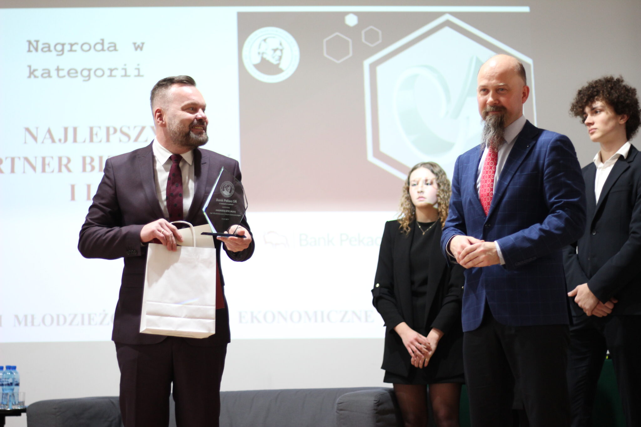 Mężczyzna w bordowym garniturze i bordowo-białej krawacie trzyma statuetkę i torbę upominkową. Obok niego prowadzący z brodą, w granatowej marynarce. W tle slajd z napisem: „Nagroda w kategorii: Najlepszy partner biznesowy I MFE” oraz logotypy szkoły i Banku Pekao.