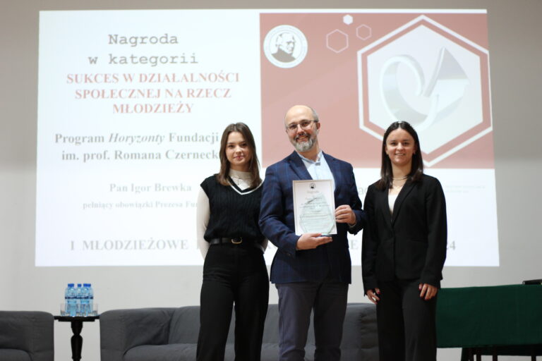 Zdjęcie przedstawia moment wręczenia nagrody podczas wydarzenia edukacyjnego „I Młodzieżowe Forum Ekonomiczne – Staszic 2024”. W centrum kadru znajdują się trzy osoby pozujące do wspólnej fotografii – mężczyzna i dwie młode kobiety. Mężczyzna trzyma dyplom z nagrodą. W tle widoczny jest duży ekran z rzutnikiem, prezentujący informacje o nagrodzie i jej laureacie. Scena znajduje się w szkolnej sali konferencyjnej. Szczegółowy opis postaci i ustawienia: Osoby: Środek kadru: Dorosły mężczyzna w średnim wieku, siwiejący, z krótkim zarostem i okularami. Ubrany w elegancką marynarkę w granatową kratę i jasnoniebieską koszulę. Trzyma w dłoniach przezroczystą ramkę lub oprawiony dyplom z tekstem i logiem szkoły. Po jego lewej stronie (prawa strona zdjęcia): Młoda kobieta z długimi, ciemnymi włosami, ubrana w czarną marynarkę i jasny top. Uśmiecha się lekko i stoi z rękami wzdłuż ciała. Po jego prawej stronie (lewa strona zdjęcia): Młoda kobieta z długimi brązowymi włosami, ubrana w czarne spodnie i czarny, dzianinowy bezrękawnik z białą koszulą z kołnierzem pod spodem. Również uśmiechnięta, ma ręce schowane za plecami. Tło: W tle znajduje się ekran projekcyjny z wyświetlonym slajdem. Slajd zawiera tekst i grafikę: Nagłówek: „Nagroda w kategorii SUKCES W DZIAŁALNOŚCI SPOŁECZNEJ NA RZECZ MŁODZIEŻY” (część w czerwonym kolorze). Opis: „Program Horyzonty Fundacji im. prof. Romana Czarneckiego” Dalej: „Pan Igor Brewka pełniący obowiązki Prezesa Fundacji” – mężczyzna na zdjęciu to najprawdopodobniej pan Igor Brewka. W prawym górnym rogu slajdu umieszczono logo I LO im. Staszica (okrągłe, czarno-białe z profilem mężczyzny) oraz czerwony sześciokąt z trójwymiarową, srebrną strzałką skierowaną w górę i prawo. Na samym dole ekranu – częściowo widoczny napis: „I MŁODZIEŻOWE FORUM EKONOMICZNE STASZIC 2024”. Scenografia sali: Przed osobami na zdjęciu stoją dwa ciemnoszare fotele i mały czarny stolik z sześcioma butelkami wody mineralnej. Po prawej stronie kadru widoczny ciemnozielony obrus przykrywający stół lub mównicę, częściowo poza kadrem. Podłoże jasne, scena dobrze oświetlona światłem sztucznym. Znaczenie zdjęcia: Fotografia dokumentuje uroczysty moment wręczenia nagrody Fundacji Horyzonty w ramach forum ekonomicznego dla młodzieży. Nagroda dotyczy działań społecznych na rzecz młodych ludzi. Zdjęcie podkreśla współpracę między instytucjami edukacyjnymi a fundacjami wspierającymi rozwój młodzieży.