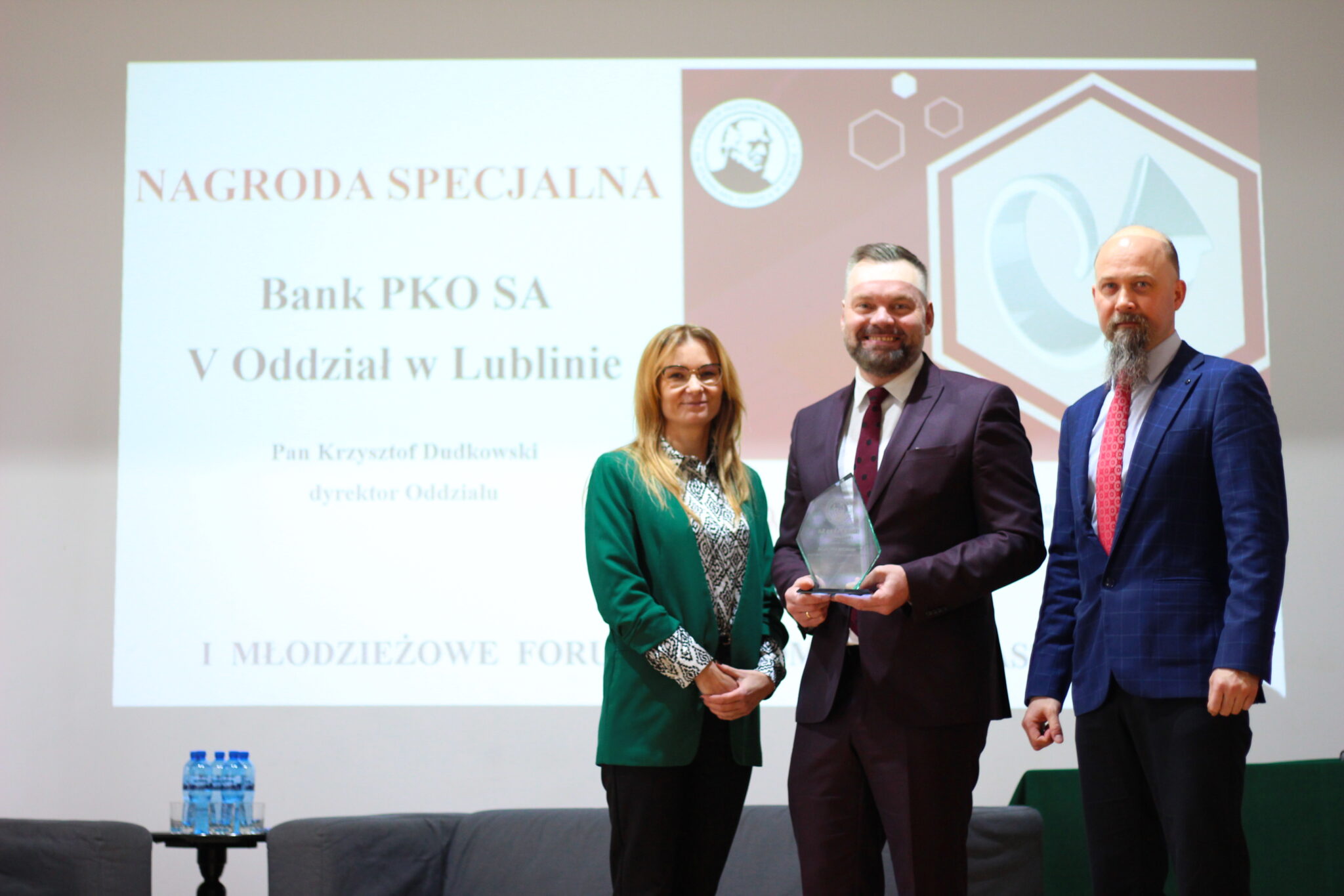 Na tle ekranu z napisem: „Nagroda Specjalna – Bank PKO SA V Oddział w Lublinie – Pan Krzysztof Dudkowski, dyrektor oddziału” stoją trzy osoby. W środku mężczyzna w bordowym garniturze trzyma przezroczystą statuetkę. Po jego lewej stronie (dla widza: prawa) stoi mężczyzna z siwą brodą w granatowej marynarce z czerwoną krawatą. Po prawej stronie kobieta w zielonej marynarce i okularach. W tle czerwono-białe logo forum i symbole graficzne.