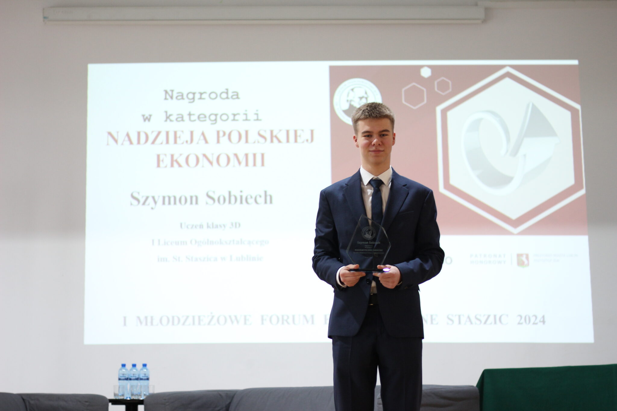 Młody chłopak w granatowym garniturze i krawacie pozuje na tle prezentacji z napisem: „Nagroda w kategorii: Nadzieja Polskiej Ekonomii – Szymon Sobiech, uczeń klasy 3D, I LO im. Staszica w Lublinie”. W rękach trzyma nagrodę – statuetkę z przezroczystego szkła. Stoi centralnie na tle sceny.