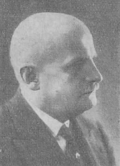 Czarno-biała fotografia mężczyzny w średnim wieku ukazanego w profilu. Ma ciemne, zaczesane do tyłu włosy oraz wyraźny wąs. Ubrany jest elegancko w ciemny garnitur, kamizelkę i krawat. Tło neutralne, portret formalny, prawdopodobnie z początku XX wieku. Twarz spokojna, skupiona.