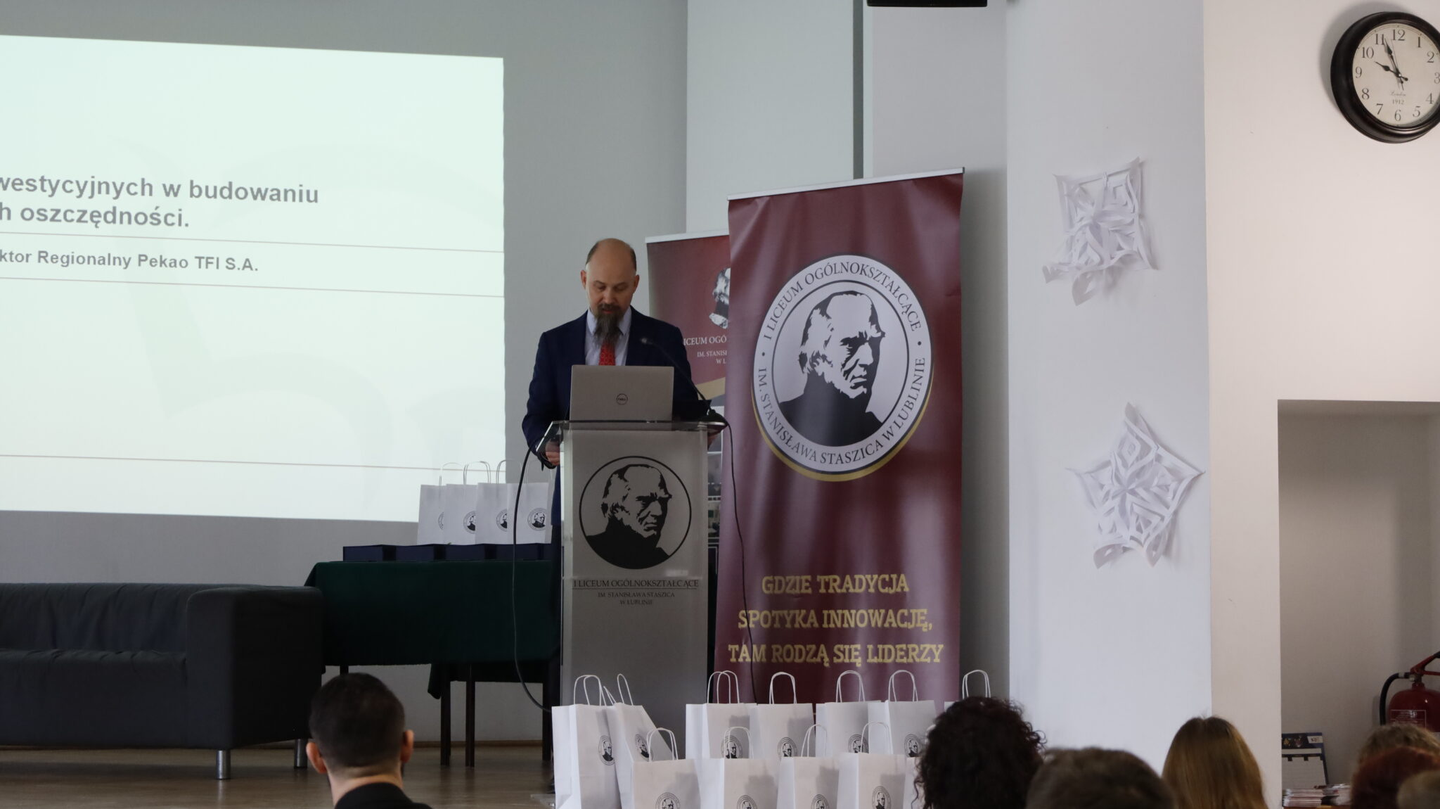 Mężczyzna z brodą w granatowym garniturze i czerwonej krawacie stoi przy przezroczystej mównicy z logo szkoły. W tle slajd prezentacji pt. „Rola funduszy inwestycyjnych w budowaniu długoterminowych oszczędności”, podpisany przez Dyrektora Regionalnego Pekao TFI S.A. Wokół mównicy stoją torby upominkowe z logotypem szkoły.