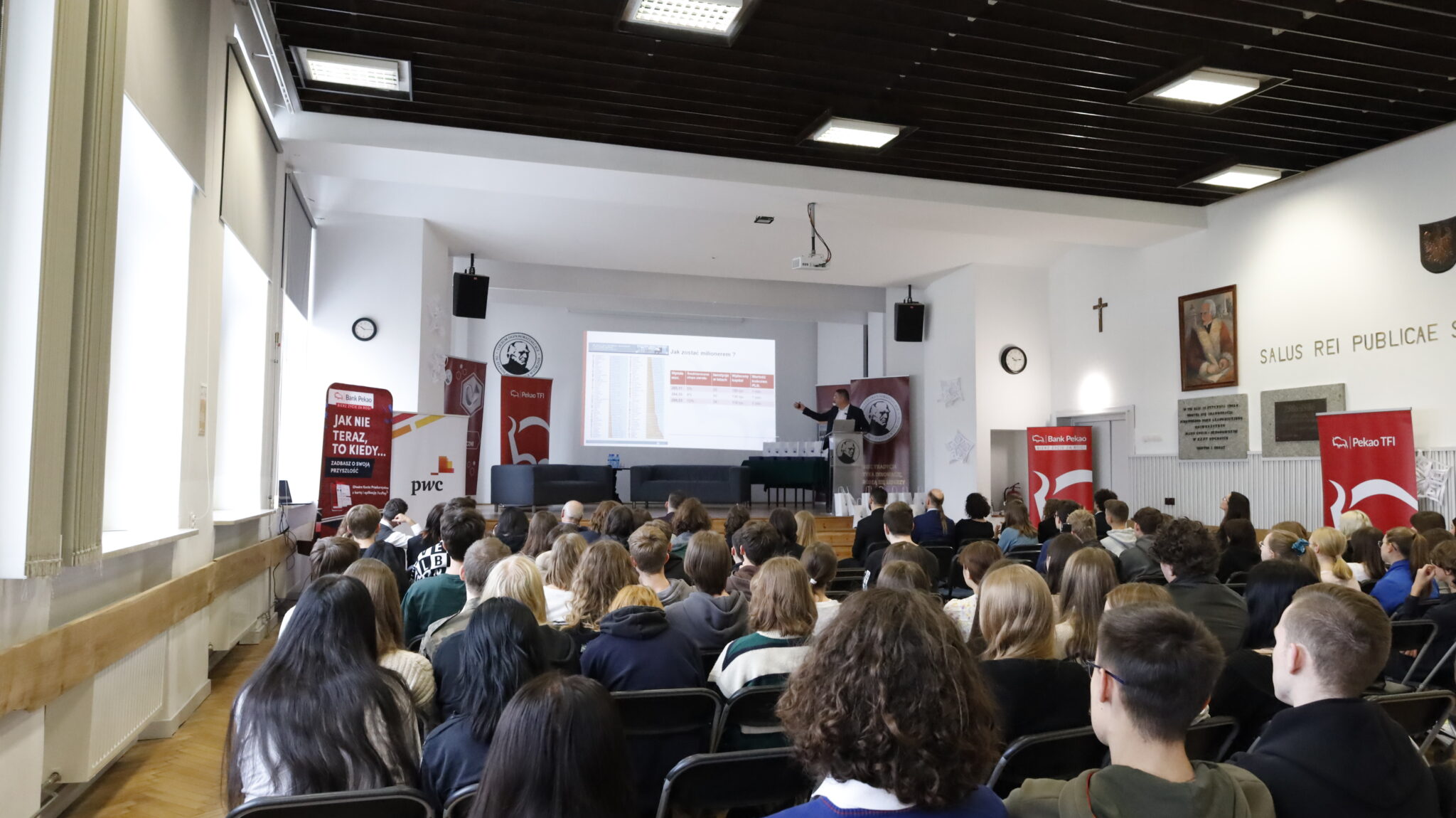Ujęcie szerokie całej sali. Przed dużym ekranem z prezentacją siedzi liczna grupa uczniów, skierowana twarzami w stronę mównicy. Prezentacja na ekranie dotyczy tematu: „Jak zostać milionerem?” – widoczna tabela z porównaniem form inwestowania. Na sali dominują kolory bieli i czerwieni – banery sponsorów (Pekao, PWC), logotypy, elementy dekoracji. Na ścianie z tyłu znajduje się krzyż i napis: „Salus rei publicae suprema lex esto” (dobro Rzeczypospolitej niech będzie najwyższym prawem).