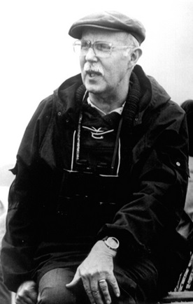 Czarno-białe zdjęcie przedstawia starszego mężczyznę w ujęciu siedzącym. Ma na głowie czapkę typu kaszkiet, nosi okulary i ma wąsy. Ubrany jest w sportową kurtkę z podniesionym kołnierzem, pod którą widać sweter i koszulę. Na szyi zawieszone ma lornetki. Jego lewa dłoń spoczywa na kolanie, a na nadgarstku nosi zegarek. Mężczyzna patrzy w bok i wygląda, jakby był w trakcie rozmowy lub tłumaczenia czegoś. Tło jest rozmyte, zdjęcie ma charakter reporterski lub dokumentacyjny.