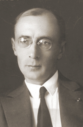 Czarno-biała fotografia portretowa przedstawia mężczyznę w średnim wieku, ujętego do ramion. Ma krótkie, starannie zaczesane włosy i nosi okulary w cienkich, okrągłych oprawkach. Jego twarz jest szczupła, o wyrazistych rysach i skupionym spojrzeniu skierowanym lekko w bok. Ubrany jest w garnitur, białą koszulę i wąski czarny krawat. Zdjęcie ma charakter formalny, wykonane zostało najprawdopodobniej w pierwszej połowie XX wieku, w stylu klasycznego portretu urzędowego lub naukowego.