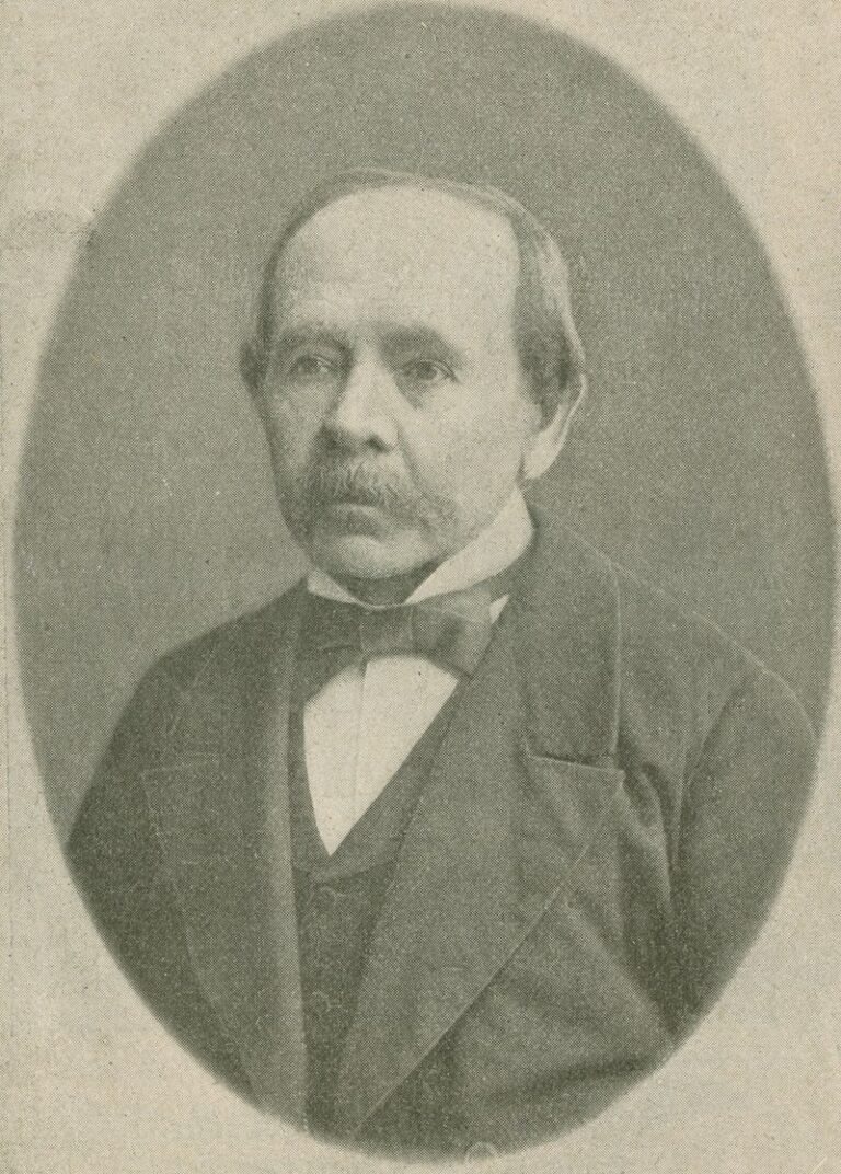 Czarno-białe, owalne zdjęcie portretowe przedstawia starszego mężczyznę, ujętego od pasa w górę. Mężczyzna ma poważny wyraz twarzy, krótkie, przygładzone włosy z zakolami i wąsy. Ubrany jest elegancko – w ciemną marynarkę, kamizelkę, białą koszulę i muszkę. Patrzy na wprost, nieco poza kadr. Tło zdjęcia jest jednolite i jasne, bez żadnych szczegółów. Styl ubioru i sposób wykonania zdjęcia sugerują koniec XIX lub początek XX wieku.