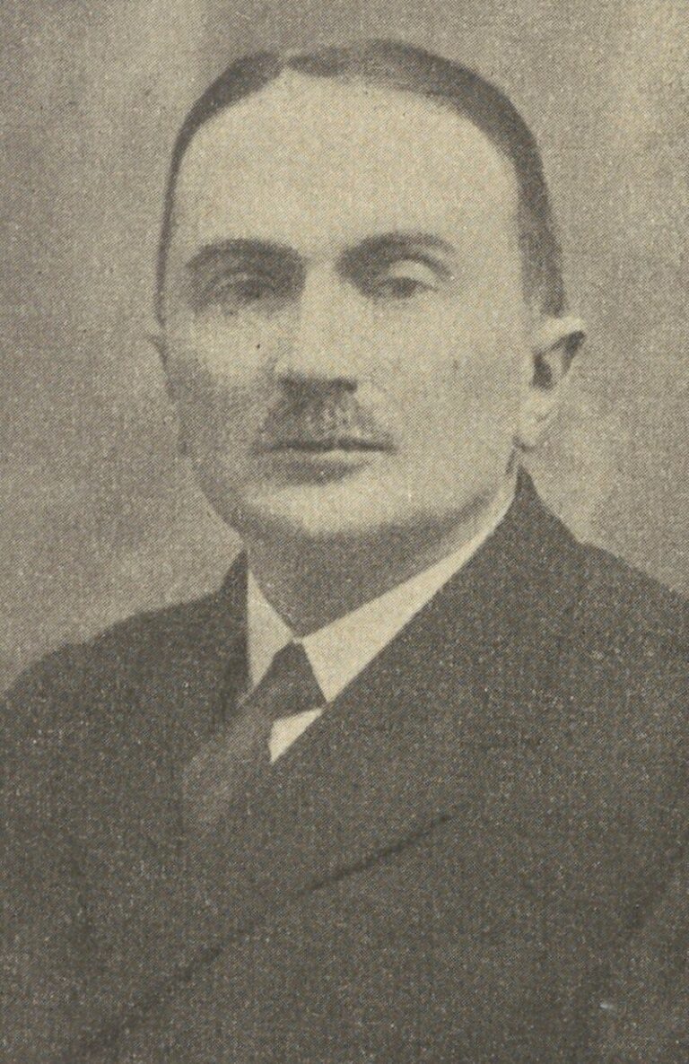 Czarno-biała, lekko ziarnista fotografia portretowa przedstawia mężczyznę w średnim wieku lub starszego, ubranego w garnitur z krawatem. Mężczyzna ma gładko zaczesane, ciemne włosy, wąski wąs i spokojny, poważny wyraz twarzy. Patrzy prosto w obiektyw. Zdjęcie ma charakter oficjalny i reprezentacyjny, wykonane najprawdopodobniej w pierwszej połowie XX wieku, na neutralnym tle.