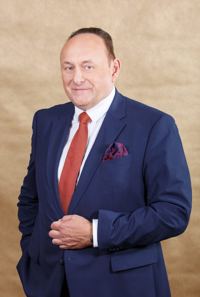 Kolorowe zdjęcie portretowe przedstawia mężczyznę w średnim wieku, stojącego na tle jasnobrązowego, jednolitego tła. Ubrany jest elegancko – ma granatowy garnitur, białą koszulę i czerwoną krawatkę w drobny wzór. W kieszonce marynarki widoczna jest ozdobna poszetka w odcieniach czerwieni i granatu. Mężczyzna ma krótko ostrzyżone, zaczesane do tyłu włosy, jasną cerę i delikatny uśmiech. Sprawia wrażenie pewnego siebie i zadowolonego. Jego lewa dłoń przytrzymuje poły marynarki na wysokości brzucha.