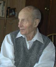 Kolorowe zdjęcie przedstawia starszego mężczyznę siedzącego w pomieszczeniu, zwróconego lekko w lewo, jakby spoglądał na coś poza kadrem. Ma rzadkie, siwe włosy i wyraźne rysy twarzy z licznymi zmarszczkami, świadczącymi o podeszłym wieku. Ubrany jest w białą koszulę z kołnierzykiem i szary, wełniany sweter bez rękawów. W tle widoczne są drewniane meble, półki z książkami i zdjęciami oraz zamknięte drzwi. Zdjęcie ma charakter domowy, niepozowane, intymne.