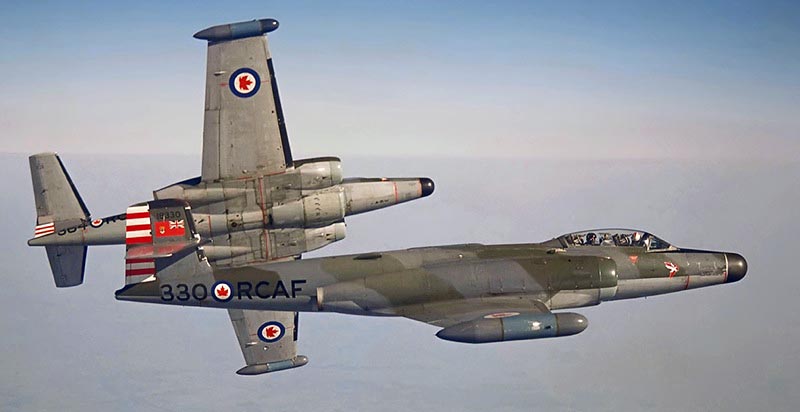 Kolorowe zdjęcie wykonane w powietrzu przedstawia dwa odrzutowce typu CF-100 Canuck w barwach Królewskich Kanadyjskich Sił Powietrznych (RCAF), lecące w szyku. Na kadłubach widoczne są oznaczenia: biało-czerwone kokardy (roundele), numer taktyczny "330" oraz napisy "RCAF". Jeden z samolotów widziany jest z boku, drugi z góry. Maszyny lecą na tle błękitnego nieba. Zdjęcie ma charakter historyczno-techniczny i upamiętnia lotnictwo wojskowe lat 50. XX wieku.