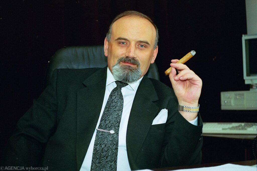 Kolorowe zdjęcie przedstawia dojrzałego mężczyznę siedzącego przy biurku w eleganckim, biurowym otoczeniu. Mężczyzna ma łysinę na czubku głowy, siwe włosy po bokach, wyraźnie zaznaczoną brodę i wąsy. Ubrany jest w ciemny garnitur, białą koszulę i ozdobny krawat z wzorem, a w kieszonce marynarki widoczna jest biała poszetka. Na nadgarstku nosi zegarek. Mężczyzna z lekkim uśmiechem trzyma w prawej dłoni zapalonego cygara. W tle widoczny jest stary komputer i monitor – co sugeruje, że zdjęcie wykonano w latach 90. XX wieku. Zdjęcie ma charakter formalny, ale z nieco swobodną, pewną siebie pozą.