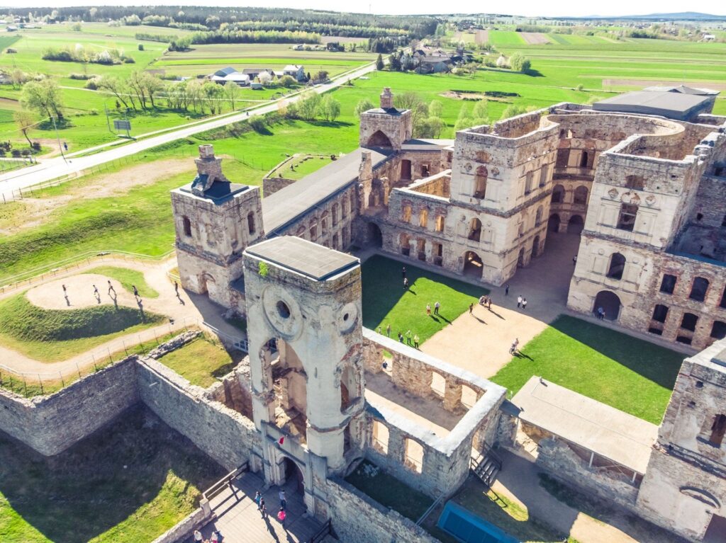 Zdjęcie przedstawia rozległe ruiny monumentalnego zamku Krzyżtopór w Ujeździe (województwo świętokrzyskie, Polska), wykonane z lotu ptaka przy pięknej, słonecznej pogodzie. Cała scena ukazana jest z dużą ilością szczegółów – zamek znajduje się na środku kadru i dominuje nad zielonym, wiejskim krajobrazem w tle.
Opis głównych elementów zdjęcia:
1. Architektura i ruiny zamku:Forma: Zamek ma kształt czworoboku z dużym, pustym dziedzińcem w środku. Mury są częściowo zrujnowane – brak dachów, częściowo zawalone ściany i brak górnych pięter w wielu miejscach.Wieże: Widoczne są cztery główne narożne wieże – masywne, wielopiętrowe, z prostokątnymi otworami po dawnych oknach. Najbardziej charakterystyczna jest wieża bramna – centralna w dolnej części zdjęcia – z dużym łukowatym przejazdem, dwoma okrągłymi otworami przypominającymi oczodoły i prostokątnym wejściem pod spodem. Przed tą wieżą znajduje się kładka i wejście dla zwiedzających.Dziedziniec: Wnętrze zamku to spory dziedziniec z zieloną trawą i piaszczystymi alejkami. Spacerują po nim grupki turystów – osoby są widoczne jako drobne sylwetki, rzucające cienie od ostrego słońca.2. Elementy krajobrazu wokół zamku:Zieleń: Zamek otoczony jest przez soczyście zielone łąki i pola uprawne, przecinane drogami i alejami. Na horyzoncie widoczne są pasma drzew, lasy oraz pola w różnych odcieniach zieleni i brązu – świadczące o wiosennej lub letniej porze roku.Drogi i zabudowania: W górnej części zdjęcia, poza murami zamku, biegnie asfaltowa droga z pojedynczymi samochodami. Poza nią rozciąga się wieś z niskimi domami i gospodarstwami, zbudowanymi głównie z jasnych materiałów, z dachami w różnych kolorach.Ruch turystyczny: Widać osoby spacerujące zarówno w obrębie zamku, jak i na alejce prowadzącej do wejścia. Są to turyści, niektórzy robią zdjęcia, inni chodzą w grupkach.3. Warunki oświetleniowe:Słońce: Scena jest bardzo dobrze oświetlona – słońce znajduje się wysoko, niebo jest bezchmurne. Cienie są ostre, co świadczy o południowej porze dnia.Kolory: Dominują jasne, naturalne kolory – beże i szarości ruin, zieleń trawy i pól, błękit nieba.Podsumowanie charakterystyki zamku:Krzyżtopór to potężna XVII-wieczna rezydencja obronna o barokowej formie. Mimo stanu ruiny, zamek imponuje rozmiarami, symetrią oraz rozmachem architektonicznym – z monumentalnymi wieżami, arkadowymi krużgankami i otwartym dziedzińcem. Współcześnie jest atrakcją turystyczną, częściowo zabezpieczoną dla zwiedzających.