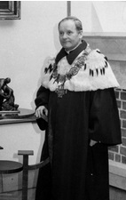 Czarno-białe zdjęcie przedstawia mężczyznę w stroju rektorskim, stojącego we wnętrzu reprezentacyjnej sali – obok widoczne są fragmenty kominka, obraz i rzeźba. Mężczyzna ma krótkie, zaczesane włosy, spokojny wyraz twarzy i delikatny uśmiech. Ubrany jest w ceremonialny, długi płaszcz rektorski z jasnym futrzanym kołnierzem i ozdobnym łańcuchem z dużym medalionem. Stoi oparty o barierkę, z ręką wspartą na niewysokim meblu. Zdjęcie ma charakter oficjalny i najprawdopodobniej pochodzi z drugiej połowy XX wieku.