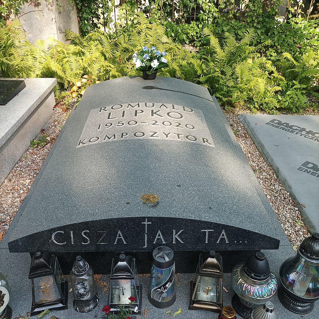 Kolorowe zdjęcie przedstawiające nowoczesny nagrobek Romualda Lipko. Na płycie z ciemnego granitu wyryto napis „ROMUALD LIPKO 1950–2020 KOMPOZYTOR”, a poniżej łukowy napis: „CISZA JAK TA…”. Przed nagrobkiem ustawiono liczne znicze w różnych kształtach i kolorach, a nad płytą znajduje się bukiet sztucznych kwiatów. Grób otacza bujna roślinność i żwir, co nadaje mu spokojny i estetyczny charakter.
