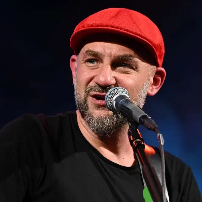 Na zdjęciu znajduje się dorosły mężczyzna występujący na scenie. Ma na sobie czerwoną, materiałową czapkę typu kaszkiet, która szczelnie przylega do głowy. Mężczyzna ma krótko przycięty, siwiejący zarost – brodę i wąsy. Jego cera jest jasna. Ubrany jest w prostą, czarną koszulkę z krótkim rękawem. Trzyma gitarę, a jego ręka opiera się na pasku od instrumentu. Śpiewa lub mówi do mikrofonu – widoczny jest mikrofon sceniczny na czarnej statywie, umieszczony blisko jego ust. Mężczyzna patrzy w lewo (z perspektywy widza). Tło jest jednolicie ciemne – odcienie granatu i czerni – z efektem dymu lub mgły scenicznej, typowym dla oświetlenia koncertowego. Zdjęcie zrobione z bliska, wyraźnie widoczne są rysy twarzy.