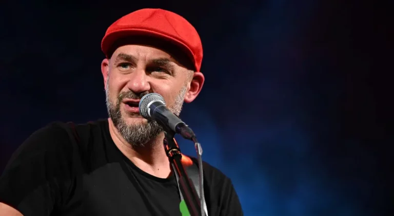 Na zdjęciu znajduje się dorosły mężczyzna występujący na scenie. Ma na sobie czerwoną, materiałową czapkę typu kaszkiet, która szczelnie przylega do głowy. Mężczyzna ma krótko przycięty, siwiejący zarost – brodę i wąsy. Jego cera jest jasna. Ubrany jest w prostą, czarną koszulkę z krótkim rękawem. Trzyma gitarę, a jego ręka opiera się na pasku od instrumentu. Śpiewa lub mówi do mikrofonu – widoczny jest mikrofon sceniczny na czarnej statywie, umieszczony blisko jego ust. Mężczyzna patrzy w lewo (z perspektywy widza). Tło jest jednolicie ciemne – odcienie granatu i czerni – z efektem dymu lub mgły scenicznej, typowym dla oświetlenia koncertowego. Zdjęcie zrobione z bliska, wyraźnie widoczne są rysy twarzy.