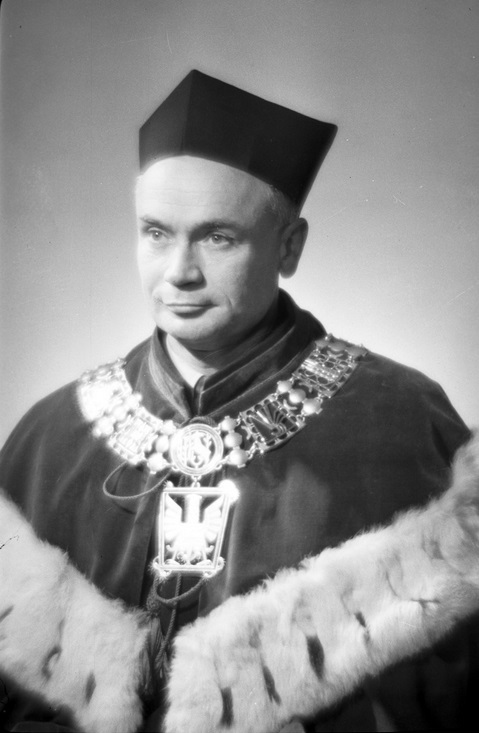 Czarno-białe, studyjne zdjęcie portretowe przedstawia mężczyznę w stroju rektorskim. Ma krótko ostrzyżone włosy i spokojny, poważny wyraz twarzy. Ubrany jest w aksamitny, ceremonialny płaszcz z szerokim obszyciem z jasnego futra oraz nosi łańcuch rektorski z dużym medalionem z orłem i symbolicznymi zdobieniami. Na głowie ma tradycyjną czapkę rektorską (biret). Mężczyzna patrzy lekko w bok. Zdjęcie ma oficjalny, uroczysty charakter i pochodzi prawdopodobnie z połowy XX wieku.