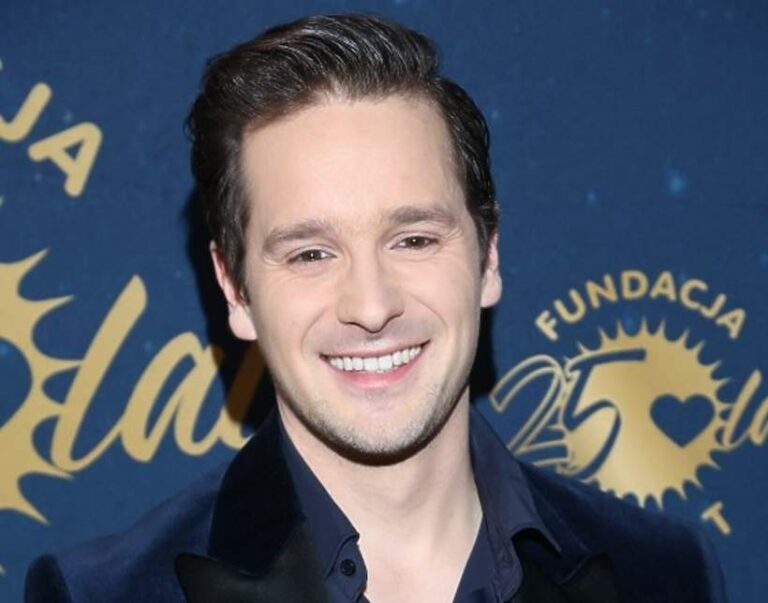 Na zdjęciu znajduje się młody mężczyzna stojący na tle ciemnoniebieskiej ścianki z napisem „Fundacja” i złotym logo w kształcie promienistego słońca. Wewnątrz logo widnieje liczba „25” oraz symbol serca. Mężczyzna ma jasną cerę i gęste, ciemne włosy zaczesane do tyłu. Uśmiecha się szeroko, pokazując zęby. Ma na sobie elegancką marynarkę z ciemnego aksamitu oraz ciemnoniebieską koszulę. Tło sugeruje, że zdjęcie zostało wykonane podczas oficjalnego wydarzenia lub gali charytatywnej. Emocje i kontekst: Mężczyzna wygląda na zadowolonego, pewnego siebie i odprężonego. Pozytywna atmosfera wydarzenia sugeruje jego celebracyjny charakter.