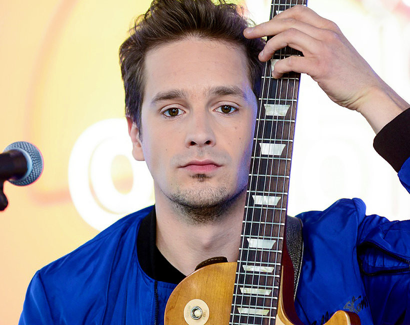 Opis: Na zdjęciu mężczyzna pozuje z gitarą. Gitara klasyczna elektryczna typu Les Paul – w odcieniach jasnobrązowego drewna – oparta jest pionowo o jego twarz. Lewą dłonią trzyma gryf gitary nad głową, a jego palce lekko oplatają instrument. Ma poważny, skupiony wyraz twarzy – nie uśmiecha się. Ubrany jest w jaskrawoniebieską, sportową kurtkę bomberkę z czarnym kołnierzykiem. W tle rozmazany, jasnożółty logotyp oraz mikrofon z lewej strony kadru sugerują, że znajduje się na scenie lub podczas prób muzycznych. Emocje i kontekst: Mężczyzna prezentuje się poważnie i skoncentrowanie. Zdjęcie podkreśla jego muzyczne zainteresowania i artystyczną ekspresję.