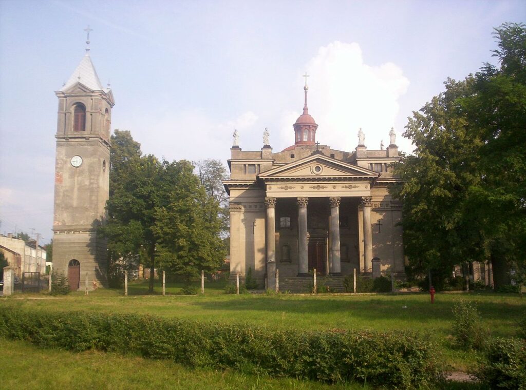 Zdjęcie przedstawia zabytkowy, monumentalny kościół z przylegającą do niego dzwonnicą. Obiekt znajduje się w otoczeniu zieleni – widać drzewa i przycięty trawnik z niskim żywopłotem w pierwszym planie.
Główna bryła kościoła:Styl architektoniczny: Klasycystyczny z elementami neorenesansu.Fasada: Symetryczna, zwrócona frontem do obserwatora. Centralną część fasady zdobi monumentalny portyk wsparty na czterech wysokich, kolumnach o gładkich trzonach w porządku korynckim. Nad kolumnami znajduje się trójkątny fronton (przypominający grecki tympanon), wewnątrz którego umieszczono okrągły otwór.Wejście główne: Znajduje się na środku, prowadzą do niego kamienne schody. Drzwi są masywne, drewniane.Nad portykiem: Widoczne są cztery rzeźby przedstawiające postacie świętych, ustawione na dachu kościoła, każda na osobnym cokole. W tle nad nimi wznosi się czerwona kopułka z małą latarnią i krzyżem na szczycie.Ściany boczne: Proste, tynkowane na jasno-szary kolor, z delikatnymi pionowymi zdobieniami imitującymi pilastry.Dzwonnica:Położenie: Po lewej stronie kościoła, oddzielona od głównego budynku.Wygląd: Czworoboczna wieża zakończona jasnym, metalowym dachem w kształcie ostrosłupa. Na szczycie krzyż.Zegar: Umieszczony w górnej części jednej ze ścian wieży. Zegar jest okrągły, z białą tarczą i czarnymi wskazówkami oraz cyframi rzymskimi.Wejście do wieży: Poniżej znajduje się proste, półokrągłe drzwi z ciemnego drewna.Otoczenie:Roślinność: Przed kościołem rozpościera się pas przystrzyżonego trawnika oraz niskie krzewy. Po obu stronach rosną drzewa liściaste, częściowo zasłaniające budynek.Pogoda i oświetlenie: Dzień jest słoneczny, niebo częściowo zachmurzone, a światło słoneczne pada z prawej strony, rzucając delikatne cienie.Ogólna atmosfera:Budynek robi wrażenie powagi i majestatu. Może być zamknięty lub opuszczony, sądząc po lekko zniszczonej elewacji (plamy, odpadający tynk) i braku ludzi. Zieleń wokół dodaje spokoju i wrażenia przestrzeni.