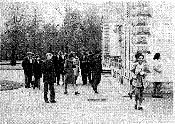 Czarno-biała fotografia, wykonana prawdopodobnie w latach 60. lub 70. XX wieku. Styl ubioru i jakość zdjęcia sugerują czas PRL-u. Zdjęcie zostało wykonane na zewnątrz, przy dużym budynku z ozdobną elewacją. Lokalizacja: Zdjęcie przedstawia grupę młodych ludzi (najprawdopodobniej studentów lub uczniów) przed dużym, reprezentacyjnym budynkiem z wyraźnie zaznaczoną architekturą klasycystyczną – widoczne są masywne boniowania (ozdobne pasy) na elewacji i balustrady z ozdobnymi poręczami. Tło i otoczenie: W tle widać drzewa liściaste, prawdopodobnie park lub teren zielony przy uczelni lub instytucji publicznej. Drzewa są bez liści – może to sugerować późną jesień albo wczesną wiosnę. Chodnik jest czysty, nie ma śniegu. Osoby na zdjęciu: Na zdjęciu widoczna jest grupa około 20 osób, w większości młodych dorosłych, idących po chodniku od lewej do prawej strony kadru. Osoby są ubrane w jesienne lub wiosenne płaszcze, większość z nich to kobiety w spódnicach lub sukienkach do kolan, mężczyźni w ciemnych kurtkach i płaszczach. Niektóre kobiety mają na sobie płaszcze z paskami, inne trzymają torebki lub zeszyty. Z przodu kadru, po prawej stronie, idą dwie kobiety – jedna z nich uśmiechnięta, niesie zeszyty i książki przytulone do klatki piersiowej, ubrana jest w jasny płaszczyk z dużymi guzikami i krótką spódniczkę, a na nogach ma ciemne buty. Obok niej idzie druga kobieta, lekko cofnięta w kadrze, ubrana w biały płaszcz – stoi tyłem do aparatu i patrzy na ścianę budynku. Grupa z tyłu wydaje się poruszać niespiesznym krokiem, niektórzy rozmawiają ze sobą, inni patrzą pod nogi, jakby wracali ze spaceru lub zajęć. Panuje spokojna, codzienna atmosfera. Kompozycja: Zdjęcie ma lekko ukośną perspektywę – budynek znajduje się po prawej stronie zdjęcia, a ludzie idą po chodniku z lewej strony w kierunku prawego przodu kadru. Główna grupa ludzi jest w centrum kadru, ale najbardziej wyróżniają się dwie kobiety z przodu – jedna idąca wprost na aparat z uśmiechem (tworzy silny punkt przyciągający uwagę). Emocje i klimat: Zdjęcie oddaje spokojną atmosferę codzienności – prawdopodobnie przerwa między zajęciami, wyjście z lekcji lub rozpoczęcie dnia. Brak pośpiechu, raczej refleksyjny nastrój. Brak nowoczesnych elementów (telefonów, plecaków itp.) potęguje wrażenie przeszłości.