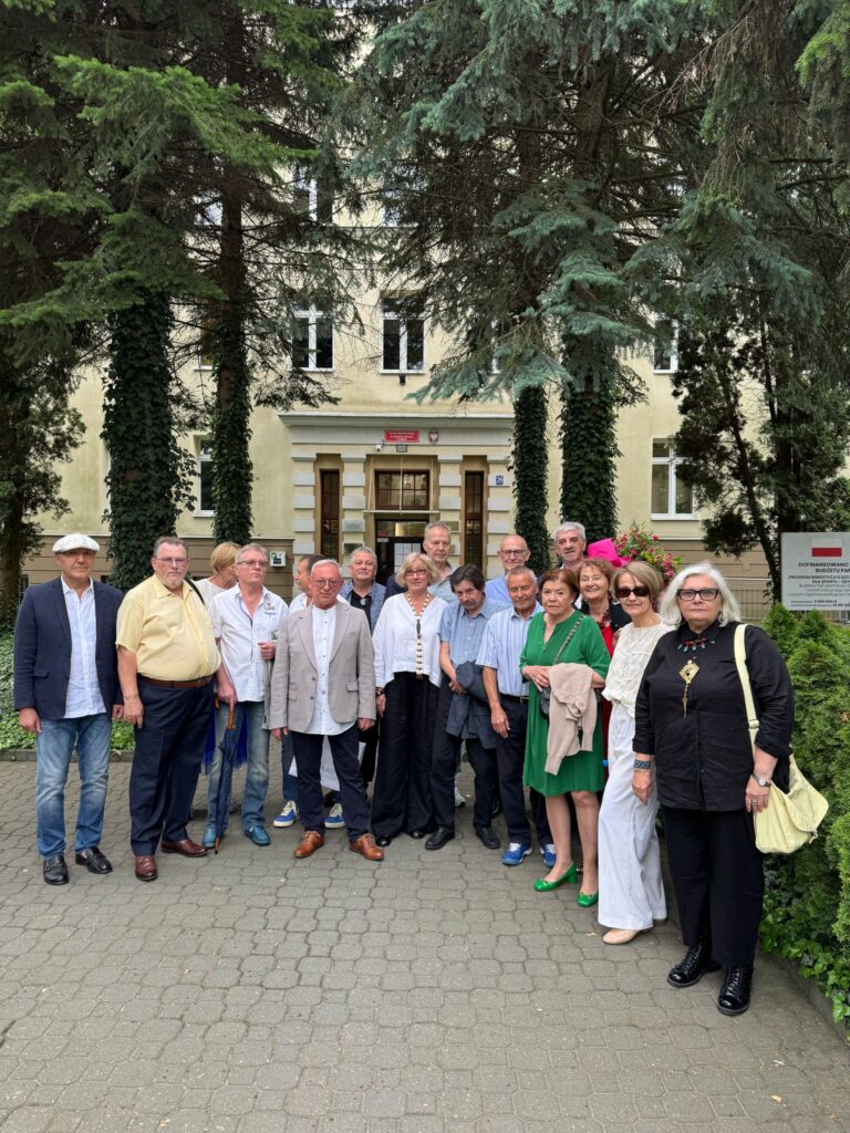 Na zdjęciu znajduje się grupa około 17 dorosłych osób (kobiety i mężczyźni) pozujących do wspólnego zdjęcia grupowego przed wejściem do budynku. Wszyscy stoją na chodniku z kostki brukowej, otoczeni wysokimi drzewami iglastymi (prawdopodobnie świerki lub jodły), które częściowo zasłaniają widok na elewację budynku. Tło i otoczenie: Budynek z jasnobeżową elewacją i białymi, dekoracyjnymi obramowaniami wokół drzwi i okien. W tle widoczny jest prostokątny, dwuskrzydłowy drewniany portal wejściowy z przeszkloną górną częścią. Nad wejściem znajduje się czerwona tablica z nazwą instytucji (niewyraźna) oraz godło Polski – biały orzeł na czerwonym tle. W lewym górnym rogu widoczne są okna. Po prawej stronie kadru – słup z tablicą informacyjną (częściowo przysłonięta krzewem). Postacie: Osoby w różnym wieku, głównie w wieku emerytalnym, ubrane w stroje eleganckie, półformalnie – koszule, marynarki, sukienki, spodnie materiałowe, buty skórzane, niektóre kobiety w kolorowych strojach (np. zielona sukienka, białe spodnie, czerwona szminka). Jedna kobieta nosi duże, ciemne okulary przeciwsłoneczne, trzyma torebkę na ramieniu. W grupie panuje pogodna atmosfera, niektóre osoby się uśmiechają. Jedna osoba ma parasol, inna ma czapkę typu kaszkiet.