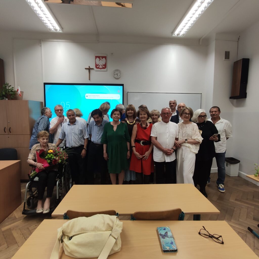 Na zdjęciu widzimy tę samą grupę (około 18 osób) sfotografowaną wewnątrz klasy szkolnej. W centrum znajduje się starsza kobieta na wózku inwalidzkim, trzymająca duży bukiet kwiatów owinięty czerwonym papierem. Pozostałe osoby stoją wokół niej, tworząc półokrąg.Tło i wnętrze:
Pomieszczenie ma białe ściany i sufit, na ścianie wisi nowoczesny ekran dotykowy z niebiesko-turkusowym tłem pulpitu (prawdopodobnie monitor interaktywny). Nad ekranem wisi godło Polski (biały orzeł) i krzyż. Widoczny jest również zegar ścienny wskazujący godzinę około 16:05. Po bokach sali znajdują się szafy, regały z roślinami oraz tablica suchościeralna.Postacie:
Uczestnicy stoją blisko siebie. Wiele osób nosi te same ubrania co na zdjęciu pierwszym – np. kobieta w czerwonej sukience, kobieta w białych szerokich spodniach i białej bluzce, mężczyzna w białej koszuli z krótkim rękawem. W tle widać kobietę o siwych włosach w czerni oraz kilku mężczyzn w koszulach. Przed pierwszymi ławkami leży beżowa torebka oraz leżą okulary i telefon komórkowy w niebieskim etui z mapą.Atmosfera:
Zdjęcie dokumentuje spotkanie towarzyskie lub okolicznościowe, prawdopodobnie z okazji jubileuszu, zjazdu klasowego lub pożegnania nauczyciela. Wyraźnie widoczna jest serdeczna, przyjazna atmosfera. Niektóre osoby się uśmiechają, inne patrzą poważnie w stronę obiektywu.