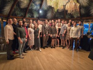 Na zdjęciu znajduje się grupa 30 młodych dorosłych osób, stojących wspólnie i pozujących do zdjęcia grupowego w ciepłym, przytulnym wnętrzu restauracyjnym z drewnianą podłogą i egzotyczną dekoracją. Zdjęcie wykonano wieczorem. Układ i sceneria: Grupa ustawiona jest w półokręgu – osoby z przodu mają więcej miejsca, tylne rzędy są lekko zbliżone. Zdjęcie wykonano w restauracyjnym wnętrzu z egzotycznym wystrojem – sufit pokryty jest materiałem z nadrukiem zielonych liści palm, a z góry zwisają naturalne, plecione lampy w stylu boho z wplecionymi zielonymi roślinami. W tle widoczne są duże okna, za którymi panuje zmrok – widać nieco niebieskiego wieczornego światła oraz uliczne neony lub światła. Ubrania i wygląd: Osoby są ubrane nieformalnie, ale schludnie, jak na letnie przyjęcie w restauracji: Kobiety noszą głównie lekkie sukienki (w kwiaty, wzory lub jednolite), w stonowanych kolorach. Mężczyźni mają na sobie koszule, t-shirty lub polówki – najczęściej jasne kolory, niektórzy w spodniach długich, inni w krótkich (szortach). Wśród butów przeważają sportowe: sneakersy, trampki lub sandały. Jedna osoba (mężczyzna w centrum zdjęcia) ubrana jest w pełny wojskowy mundur galowy z odznaczeniami – ciemny garnitur wojskowy z pagonami, krawatem i czapką trzymaną w dłoni. To prawdopodobnie osoba honorowana na spotkaniu. Emocje i interakcje: Wszyscy uczestnicy patrzą w stronę aparatu i uśmiechają się, tworząc przyjazną i zgraną atmosferę. Osoby stoją blisko siebie, niektóre mają ręce na plecach kolegów lub koleżanek, co podkreśla ich koleżeńską relację. Dodatkowe elementy: W prawym dolnym rogu częściowo widoczny jest ciemny fotel i fragment stolika z nakryciem. W tle na stole widoczne są szklane karafki, naczynia oraz kwiatowa dekoracja, co wskazuje, że jest to przyjęcie lub spotkanie integracyjne. Podsumowanie: Zdjęcie przedstawia dużą, zgraną grupę młodych ludzi podczas towarzyskiego spotkania w egzotycznie urządzonej restauracji wieczorem. Centralną postacią może być mężczyzna w mundurze wojskowym. Atmosfera jest radosna i serdeczna, a aranżacja wnętrza – ciepła, pełna naturalnych akcentów.