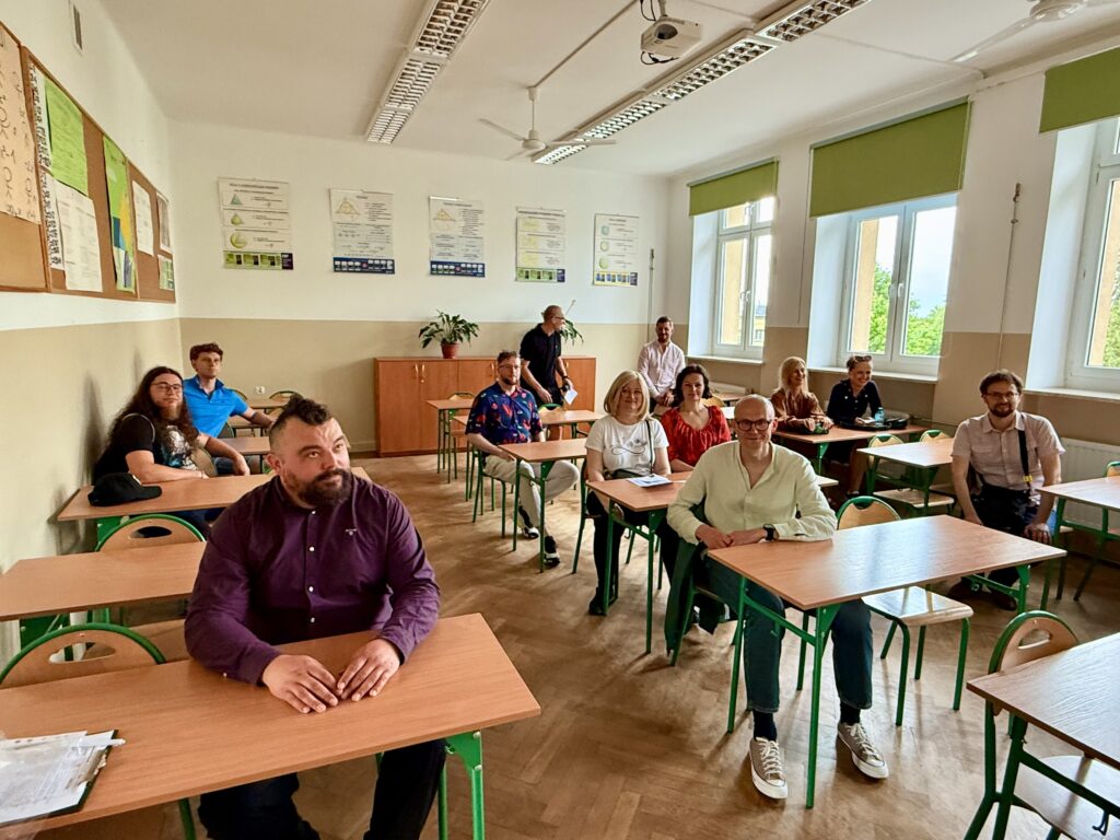 Grupa 14 osób siedzi w sali lekcyjnej przy klasycznych szkolnych ławkach z zielonymi metalowymi nogami. Ławki ustawione są w kilku rzędach, a osoby siedzą przodem do fotografa. Większość uśmiechnięta, niektóre osoby patrzą prosto w obiektyw, inne lekko na bok.Wystrój sali:Ściany biało-beżowe, dolna część ścian w jaśniejszym odcieniu beżu.Duże okna po prawej stronie wpuszczają naturalne światło. Zielone rolety są częściowo podniesione.Na ścianie z lewej strony widoczne są plakaty edukacyjne, prawdopodobnie z zakresu biologii lub chemii – zawierają wykresy i schematy.Na końcu sali szafa z doniczką i nauczyciel stojący w głębi (jedyny stojący mężczyzna).Uczestnicy:Mężczyzna z irokezem siedzi w pierwszym rzędzie po lewej stronie, w ciemnej koszuli, z rękami na stole.Kobieta z blond włosami i białą koszulką siedzi na środku – znana z poprzednich zdjęć.Osoby siedzą po dwie przy jednej ławce, rozmieszczone są równomiernie.