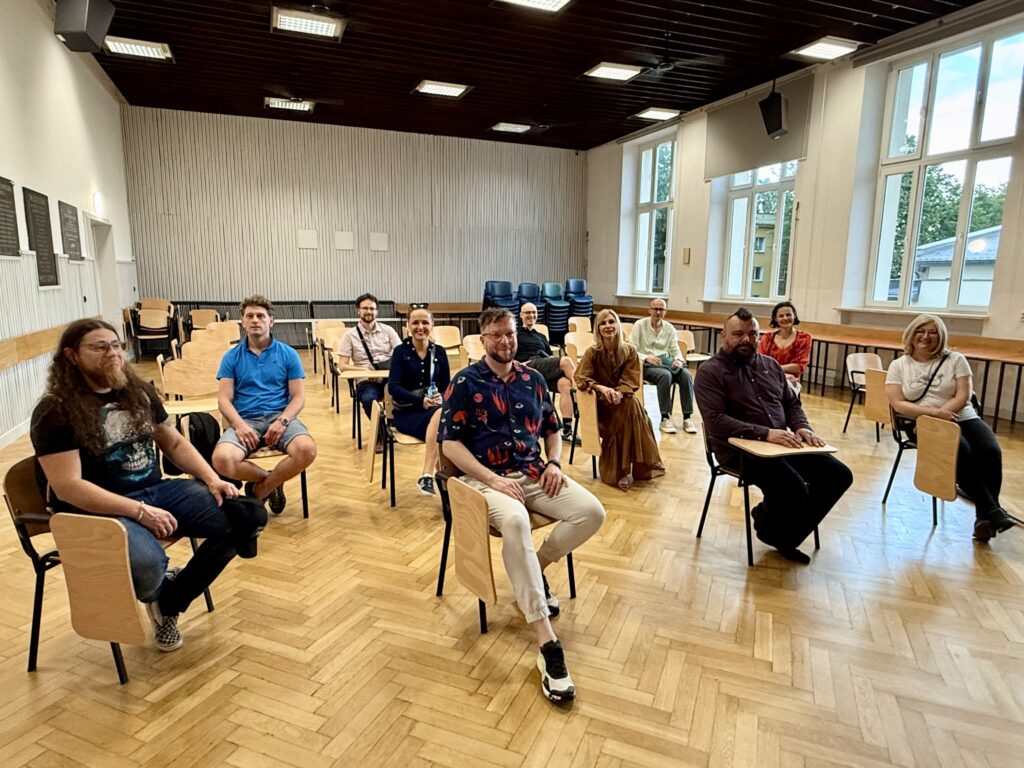 Grupa tych samych 13 osób siedzi w szkolnej sali wykładowej na krzesłach z pulpitem do pisania. Rozstawione są one w dwóch rzędach, z dużymi odstępami. Osoby skierowane są twarzami do aparatu. Pomieszczenie jest jasne, z dużymi oknami po prawej stronie i ścianą z pionową białą boazerią po lewej.Otoczenie i układ:Podłoga wykonana z parkietu w jodełkę.Krzesła są drewniane z jasnymi oparciami i siedziskami, na metalowych nogach. Pulpity przymocowane są do boków krzeseł.W głębi sali ustawione są krzesła ułożone jedno na drugim.Sufit z ciemnego drewna, z licznymi kwadratowymi panelami świetlnymi.Wyraz twarzy i atmosfera:Osoby są uśmiechnięte, zrelaksowane.Mężczyzna z długimi włosami i brodą siedzi z przodu z lewej strony, trzyma ręce na kolanach.Osoby z pierwszego zdjęcia mają te same stroje – można je łatwo zidentyfikować.