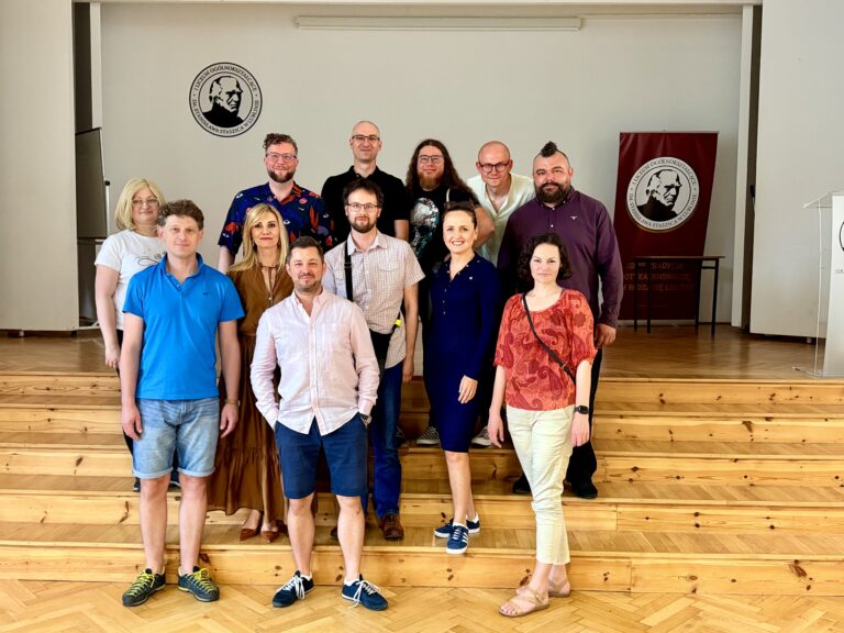 Zdjęcie przedstawia grupę 13 osób (7 mężczyzn i 6 kobiet), stojących na drewnianych schodkach sceny w szkolnej auli. Wszyscy pozują do wspólnego zdjęcia i patrzą prosto w obiektyw. Tło to jasna ściana z herbem szkoły (czarno-białe godło z wizerunkiem mężczyzny z brodą i napisem: „I Liceum Ogólnokształcące im. Stanisława Staszica w Lublinie”) oraz bordowy baner szkoły po prawej stronie. Ubiór i wygląd: W centralnym dolnym rzędzie stoi mężczyzna w różowej koszuli z krótkim rękawem i granatowych spodenkach – dłonie ma w kieszeniach, obok niego kobieta w granatowej sukience do kolan i sportowych butach, uśmiecha się szeroko. Mężczyzna z lewej w niebieskiej koszulce polo i dżinsowych spodenkach – z przodu, trzyma ręce wzdłuż ciała. Na górnych schodkach stoi mężczyzna z długimi, kręconymi włosami i brodą – ubrany w czarną koszulkę z nadrukiem, obok niego wysoki mężczyzna łysy w okularach, ubrany na czarno. Wyróżnia się mężczyzna z irokezem i brodą, ubrany w ciemnofioletową koszulę – stoi po prawej stronie, blisko banera. Inna kobieta ubrana jest w długą, brązową sukienkę, ma blond włosy i stoi z boku grupy. Otoczenie: Podłoga i schody wykonane są z lakierowanego drewna o jasnym odcieniu. Scena ustawiona jest w dużym pomieszczeniu z białymi ścianami. Po lewej stronie widoczna jest tablica lub ekran schowany w wnęce.