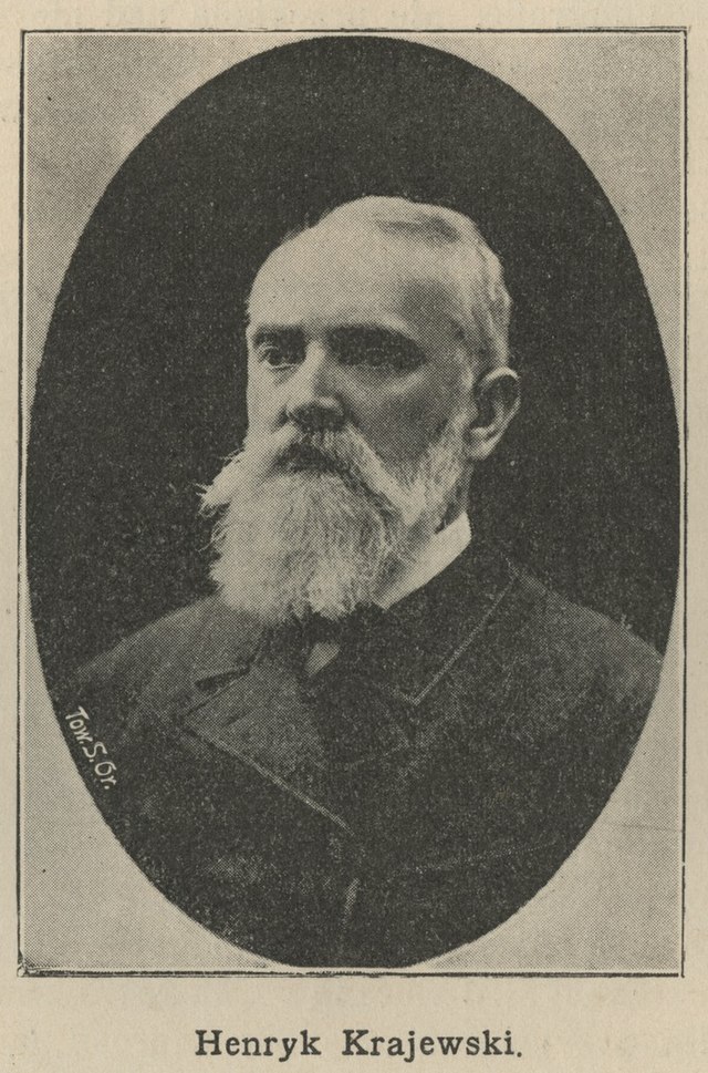 To czarno-biała fotografia w ramce w kształcie owalu. Przedstawia starszego mężczyznę z poważnym wyrazem twarzy. Mężczyzna to Henryk Krajewski, jak głosi podpis poniżej zdjęcia. Twarz prostokątna, z wysokim czołem i wyraźnie cofniętą linią włosów. Włosy krótkie, siwe, gładko uczesane. Broda długa, gęsta, biała – spływająca poniżej brody do piersi. Wąsy również długie, łączące się z brodą. Oczy głęboko osadzone, spojrzenie poważne, nieco surowe, skierowane lekko na bok. Ubrany w ciemny, elegancki garnitur z wysokim kołnierzem. Nie widać krawata ani dodatków. Zdjęcie wykonane w stylu klasycznym, atelierowym, najprawdopodobniej w końcu XIX lub na początku XX wieku.