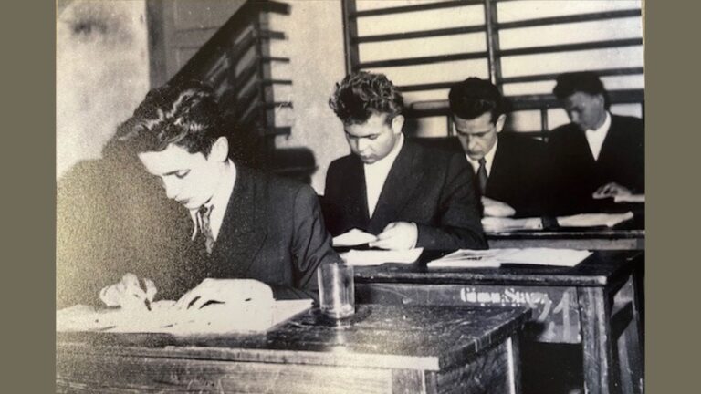 Zdjęcie czarno-białe, wykonane we wnętrzu budynku, najprawdopodobniej w klasie szkolnej lub sali egzaminacyjnej. Przedstawia czterech młodych mężczyzn siedzących w dwóch rzędach przy drewnianych ławkach, skupionych na pisaniu lub czytaniu dokumentów, prawdopodobnie podczas egzaminu lub lekcji. Ubrani są elegancko – wszyscy mają ciemne garnitury i białe koszule, trzech z nich ma także ciemne krawaty. Opis szczegółowy: Na pierwszym planie, z lewej strony zdjęcia, znajduje się młody mężczyzna z ciemnymi, gęstymi włosami, pochylony nad kartką papieru. Prawą ręką pisze, a lewą przytrzymuje kartki. Na jego ławce, z przodu, stoi przezroczysta szklanka. Bezpośrednio za nim siedzi drugi mężczyzna – ma jasne, mocno zaczesane do góry włosy. Jest pochylony nad kilkoma arkuszami papieru, które trzyma obiema rękami. Dalej, po prawej stronie kadru, siedzą jeszcze dwaj mężczyźni. Obaj również zajęci są dokumentami. Trzeci z kolei wygląda na bardzo skupionego – jego twarz skierowana jest ku dołowi, a dłonie trzymają kartki. Ostatni mężczyzna, znajdujący się najdalej w tle, jest częściowo rozmazany przez głębię ostrości zdjęcia, ale widać, że także analizuje jakieś dokumenty. W tle widoczne są elementy wystroju wnętrza – z lewej strony klatka schodowa ze schodami i barierką, z prawej typowe drabinki gimnastyczne, sugerujące, że może to być sala gimnastyczna zaadaptowana na potrzeby egzaminu. Na przedniej ściance pierwszej ławki widać namalowany numer: „71” oraz niewyraźny napis, prawdopodobnie „Gimn. Stal.” Wrażenie ogólne: Zdjęcie dokumentuje poważny, formalny moment – egzamin lub test – w czasie PRL lub w okresie powojennym, sądząc po fryzurach, ubraniach i wyposażeniu sali. Panuje skupiona atmosfera, a uczniowie wyglądają na bardzo skoncentrowanych. Całość jest w odcieniach szarości (monochromatyczna), a oświetlenie pochodzi zapewne z lamp sufitowych.