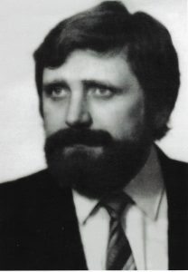 Zdjęcie czarno-białe, formalne, wykonane w stylu portretowym, prawdopodobnie w latach 70. lub 80. XX wieku. Przedstawia mężczyznę ujętego od klatki piersiowej w górę. Mężczyzna jest lekko zwrócony w lewo (z punktu widzenia widza – w prawo), ale jego głowa odwrócona jest niemal frontalnie, co nadaje portretowi oficjalny i dostojny charakter. Twarz wyraża spokój, skupienie i powagę.Wygląd mężczyzny:Mężczyzna w wieku około 40–50 lat. Ma gęste, ciemne włosy, dość długie jak na standardy portretowe – sięgające karku, z charakterystyczną falą, nieco opadającą na czoło z lewej strony. Twarz pociągła, wyrazista, z pełnym zarostem – broda i wąsy są gęste, ciemne, dobrze utrzymane, tworząc zwartą całość. Broda okala całą dolną część twarzy i łączy się z bokobrodami, co nadaje postaci poważny i lekko surowy wygląd.Oczy wyraziste, głęboko osadzone, skierowane w dal, ponad kadr – jakby mężczyzna patrzył zamyślony w przyszłość. Brwi gęste, ciemne, nos prosty i szeroki u nasady. Usta schowane częściowo pod brodą – nie widać ich wyraźnie, ale twarz wydaje się spokojna, bez uśmiechu.Ubiór:Mężczyzna ubrany jest formalnie – ma na sobie ciemny garnitur, białą koszulę i krawat w skośne pasy (kontrastowe odcienie szarości). Krawat jest szeroki, typowy dla mody z lat 70. i 80., starannie zawiązany pod klasycznym kołnierzykiem koszuli. Ubiór wskazuje na oficjalny charakter zdjęcia – może to być fotografia zawodowa, np. nauczyciela, dyrektora, pracownika urzędu lub intelektualisty.Tło i oświetlenie:Tło jednolite, jasne, bez ozdób czy tekstur – prawdopodobnie neutralna ściana fotograficzna. Światło miękkie, skierowane z przodu i lekko z lewej strony – dobrze oświetla twarz, ale pozostawia delikatne cienie, co nadaje zdjęciu głębi i charakteru.Wrażenie ogólne:Zdjęcie przedstawia osobę budzącą zaufanie, o wyrazistym wyglądzie i dostojnej prezencji. Całość kompozycji sugeruje, że mężczyzna pełnił ważną rolę w środowisku edukacyjnym lub społecznym. Portret oddaje powagę, inteligencję i spokój.