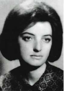 Zdjęcie czarno-białe, portretowe, wykonane w stylu typowym dla lat 60. lub 70. XX wieku. Przedstawia młodą kobietę w ujęciu od klatki piersiowej w górę, patrzącą lekko w prawo, poza kadr. Twarz kobiety jest poważna, wyraz spokojny, bez uśmiechu. Skóra jasna, owalna twarz, regularne rysy. Brwi ciemne, naturalne, oczy ciemne – wydają się być brązowe lub czarne (ze względu na brak koloru w fotografii nie można stwierdzić jednoznacznie). Nos prosty, średniej długości, usta pełne, zamknięte, w neutralnym wyrazie.Włosy są ciemne, gęste, uczesane w charakterystyczną fryzurę z dużą objętością – typowy styl kobiecy lat 60., przypominający tzw. „hełm fryzjerski”. Włosy są krótkie do ramion, gładko zaczesane, z dużą objętością na czubku głowy.Kobieta ubrana jest w ciemną bluzkę lub sukienkę z widocznym wzorem geometrycznym. Wzór to powtarzające się kwadraty, w środku których widnieje mniejszy motyw przypominający krzyż lub rozetę – tkanina wydaje się być ozdobna, z delikatnym połyskiem. Ubranie zapinane na guziki przy szyi, co widać na kołnierzyku.Tło zdjęcia jest neutralne, jasne, jednolite, bez widocznych elementów. Oświetlenie skierowane jest z przodu i lekko z góry, dzięki czemu twarz kobiety jest dobrze oświetlona, bez ostrych cieni.Zdjęcie ma charakter formalny lub dokumentacyjny – może to być fotografia do legitymacji, dokumentu lub wykonana na potrzeby publikacji. Brak ozdób, biżuterii czy elementów rozpraszających uwagę – wszystko skupia się na twarzy i fryzurze kobiety.