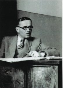 Zdjęcie czarno-białe, dokumentalne, wykonane we wnętrzu, prawdopodobnie w klasie lub gabinecie szkolnym. Przedstawia starszego mężczyznę siedzącego za drewnianym biurkiem. Zdjęcie wykonane zostało z lekkiego ukosa – postać ujęta jest od pasa w górę. Mężczyzna skierowany jest w lewo (z punktu widzenia widza – w prawo), ale jego twarz odwrócona jest w stronę rozmówcy lub fotografa, co sugeruje, że mógł być w trakcie wypowiedzi lub słuchania.Opis wyglądu mężczyzny:Mężczyzna ma około 50–60 lat, włosy ciemne, przerzedzone, zaczesane na bok, z wyraźną łysiną pośrodku. Twarz pociągła, z wyraźnymi rysami, nos prosty, usta lekko otwarte – wygląda, jakby mówił lub był w trakcie wypowiedzi. Oczy ukryte za ciemnymi, okrągłymi okularami w grubych oprawkach. Wyraz twarzy skupiony, inteligentny, zaangażowany.Ubrany jest bardzo elegancko: garnitur z jasnej tkaniny (prawdopodobnie wełniany), koszula jasna, krawat w geometryczny wzór (możliwe, że w kratę lub z dużymi rombami). Z mankietu marynarki wystaje mankiet koszuli, co sugeruje dużą dbałość o elegancję. Ubranie starannie dobrane, kojarzy się z nauczycielem, urzędnikiem lub intelektualistą z połowy XX wieku.Otoczenie i kontekst:Mężczyzna siedzi za dużym, masywnym, drewnianym biurkiem, którego front widoczny jest na pierwszym planie – drewno ma wyraźnie zarysowaną strukturę słojów. Na biurku znajdują się otwarte książki lub dokumenty, być może zeszyty – ich brzegi są nieregularne, sugerując intensywne użytkowanie. Ręka mężczyzny oparta jest na biurku – prawdopodobnie trzyma pióro lub długopis, jakby właśnie coś pisał lub podkreślał.Ściana za mężczyzną dwukolorowa: dolna część ciemniejsza, górna jaśniejsza – co sugeruje klasyczny podział malowania ścian w szkołach lub urzędach w okresie powojennym. Brak innych elementów dekoracyjnych czy mebli – skupienie na postaci i kontekście pracy umysłowej.Ogólny charakter zdjęcia:Zdjęcie ma silny charakter edukacyjny lub historyczny – przedstawia osobę, która mogła być nauczycielem, dyrektorem szkoły, naukowcem lub urzędnikiem. Kompozycja skupiona jest na atmosferze powagi, pracy intelektualnej i autorytetu. Światło miękkie, boczne, bez ostrych cieni, tworzy nastrój skupienia i refleksji.