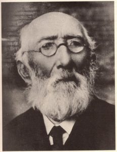 Czarno-białe zdjęcie portretowe przedstawia starszego mężczyznę, sfotografowanego od klatki piersiowej w górę. Mężczyzna patrzy lekko w lewo, poza kadr, z łagodnym, zamyślonym wyrazem twarzy. Twarz jest podłużna, z wysokim czołem i głęboko osadzonymi oczami. Mężczyzna ma bardzo rzadkie włosy na czubku głowy, natomiast po bokach głowy pozostało ich nieco więcej – są jasne, cienkie i przylegają do skóry. Charakterystycznym elementem wyglądu jest jego długa, gęsta broda – biała lub siwa – o pełnej, naturalnej strukturze, sięgająca poniżej szyi. Broda przechodzi w równie gęste wąsy, które zakrywają górną wargę. Całość nadaje twarzy dostojny, uczony wygląd. Mężczyzna nosi małe, okrągłe okulary o cienkich metalowych oprawkach. Okulary spoczywają nisko na nosie. Jego oczy są lekko przymrużone, a spojrzenie spokojne, kontemplacyjne. Nos jest prosty i wydatny. Ubrany jest elegancko: ma na sobie jasną koszulę z kołnierzykiem, ciemną marynarkę oraz ciemny krawat z wyraźnym węzłem. Styl ubioru i brody wskazuje na zdjęcie wykonane najprawdopodobniej w drugiej połowie XIX wieku lub na początku XX wieku. Tło zdjęcia jest neutralne, delikatnie rozmyte – może być to ściana lub materiał – nie zawiera żadnych szczególnych detali, co koncentruje całą uwagę na postaci. Całe zdjęcie ma charakter poważny i formalny, być może wykonane zostało z okazji jubileuszu, publikacji naukowej lub jako oficjalny portret osoby znaczącej w swoim środowisku.