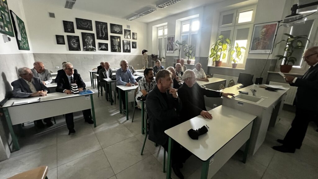 Zdjęcia przedstawiają grupę starszych mężczyzn siedzących w nowoczesnej sali lekcyjnej, urządzonej w stylu pracowni biologicznej lub medycznej. Pomieszczenie jest dobrze oświetlone światłem dziennym, a jego ściany i meble utrzymane są w jasnej tonacji. W sali znajduje się ponad piętnaście mężczyzn, w wieku około 60–80 lat, ubranych elegancko – w marynarki, garnitury, koszule. Część z nich ma siwe lub białe włosy, a kilku nosi okulary. Wszyscy siedzą przy białych ławkach z zielonymi metalowymi nogami, w równych rzędach. Na ławkach widoczne są dokumenty, zeszyty, piórniki, maska ochronna. Mężczyźni są skupieni, patrzą w jednym kierunku — na prowadzącego zajęcia lub prezentację. Panuje atmosfera uwagi i koncentracji. Tło sali: Na ścianie z lewej strony zawieszone są czarne tablice z grafikami rentgenowskimi i anatomicznymi – przedstawiają m.in. czaszkę, żebra, nerki, stopy, układ pokarmowy. W rogu sali stoi szkielet ludzki na stojaku – prawdopodobnie pomoc dydaktyczna. Przy oknach po prawej stronie znajdują się rośliny doniczkowe o dużych liściach, ustawione na parapetach, a na ścianach wiszą plakaty edukacyjne z grafikami ludzkiego ciała.