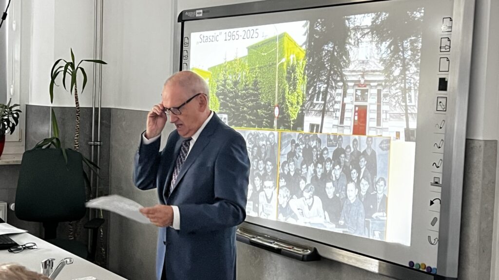 Oba zdjęcia ukazują starszego mężczyznę w granatowym garniturze, który prowadzi wykład lub prezentację w tej samej sali lekcyjnej. Mężczyzna ma siwe włosy, okulary, w jednej ręce trzyma kartkę z notatkami, a drugą unosi okulary na czoło (zdjęcie 3) lub poprawia je na nosie (zdjęcie 4). Ujęcia pokazują moment wypowiedzi, kiedy prowadzący jest w ruchu, mówi, gestykuluje, patrzy na słuchaczy. Tło prezentacji: Za nim znajduje się tablica interaktywna, na której wyświetlona jest prezentacja zatytułowana: „Staszic 1965–2025” Na ekranie widoczne są cztery fotografie: Dwa współczesne zdjęcia szkoły – zielony, obrośnięty bluszczem budynek i wejście główne z czerwonymi drzwiami. Dwa czarno-białe zdjęcia klasowe – grupowe zdjęcia uczniów sprzed kilkudziesięciu lat. Otoczenie: W tle widoczne są doniczki z roślinami oraz stanowisko nauczyciela z komputerem, kranem laboratoryjnym i innym sprzętem dydaktycznym.