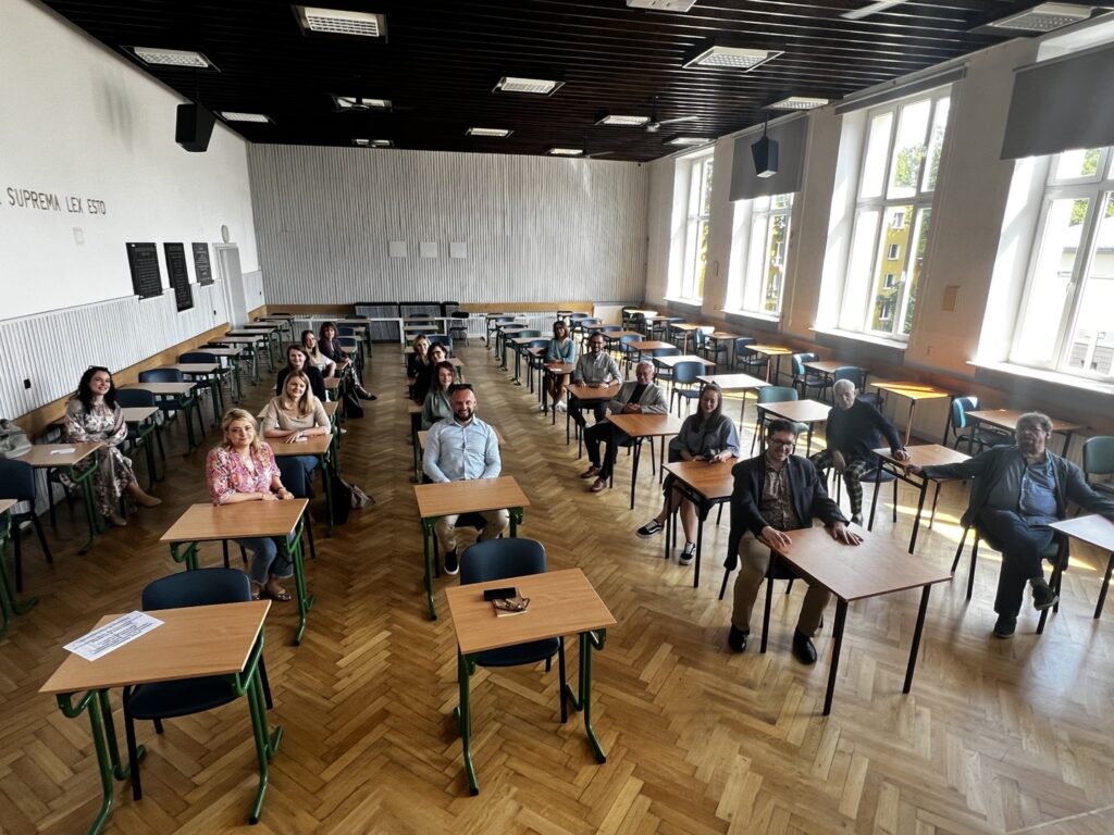 Zdjęcie przedstawia przestronną, jasną salę lekcyjną z dużymi, białymi oknami po prawej stronie. W sali ustawionych jest kilkadziesiąt szkolnych ławek z jasno-drewnianymi blatami i zielonymi metalowymi nogami oraz dopasowanymi krzesłami. W sali znajduje się 21 osób, głównie dorosłych, siedzących pojedynczo przy oddzielnych ławkach w równych rzędach, rozmieszczonych głównie w centralnej części sali. Osoby są zwrócone przodem do aparatu fotograficznego i uśmiechają się, panuje przyjazna, swobodna atmosfera. Po lewej stronie sali na ścianie widnieje fragment napisu łacińskiego: „SALUS POPULI SUPREMA LEX ESTO” – oznaczający „Dobro ludu najwyższym prawem niech będzie”. Na ścianach wiszą również czarne tablice z białymi napisami (niewyraźnymi w kadrze), prawdopodobnie z cytatami lub nazwiskami. Podłoga pokryta jest jasnym parkietem w jodełkę, sufit jest ciemny i wyłożony prostokątnymi panelami z lampami.