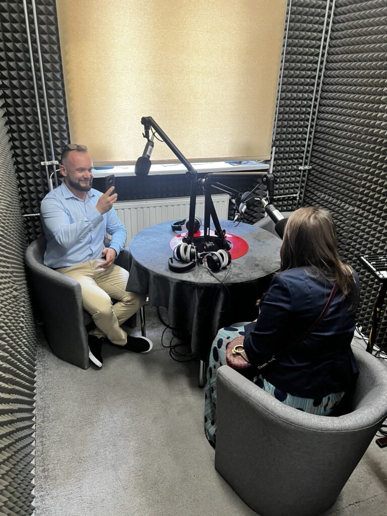 Na zdjęciu widać dwie osoby w niewielkim, profesjonalnym studiu nagraniowym przeznaczonym prawdopodobnie do nagrań audio (podcastów lub audycji). Po lewej stronie siedzi mężczyzna w niebieskiej koszuli i beżowych spodniach, uśmiechnięty, trzymający w ręce smartfona. Po prawej stronie, plecami do aparatu, siedzi kobieta z długimi brązowymi włosami. Ubrana jest w granatową marynarkę i wzorzystą spódnicę z niebieskimi kropkami, trzyma na kolanach bordową torebkę z paskiem. Między nimi stoi okrągły stół pokryty czarnym obrusem, na którym znajdują się: Dwa profesjonalne mikrofony na wysięgnikach, Para słuchawek nausznych, Przewody i inne akcesoria audio. Pomieszczenie jest wyłożone ciemnymi, piankowymi panelami wygłuszającymi na ścianach i drzwiach, a okno na wprost zasłonięte jest grubą, kremową roletą.